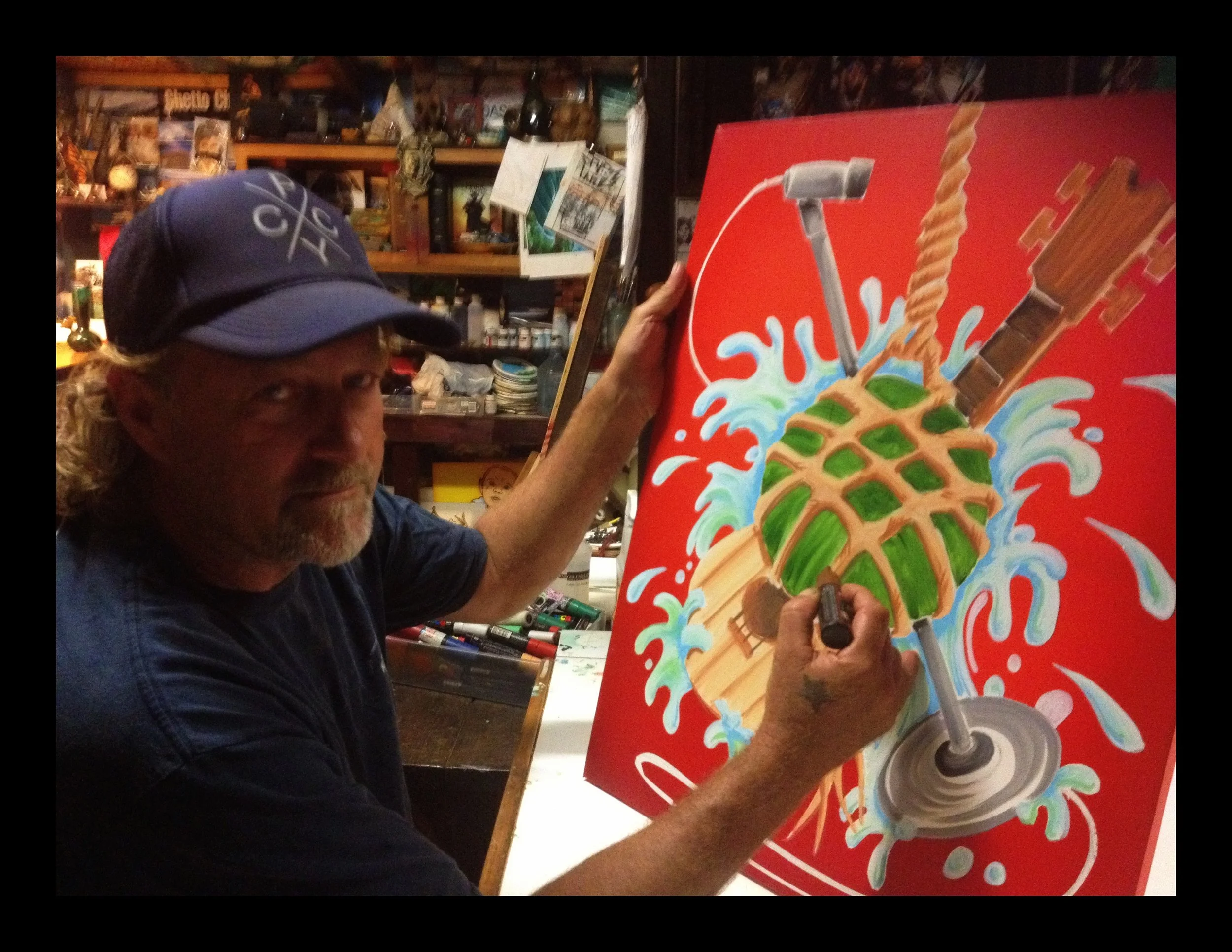 John Glomb with long curly hair and a blue baseball cap is painting a colorful piece of art featuring a pineapple combined with a drink umbrella, straws, and a fork, with splashes of water on a red background in an art studio.