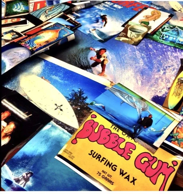 Collection of surfing-themed posters and magazine pages with vibrant images of surfers and ocean scenes, including a prominent yellow poster for Bubble Gum Surf Wax.