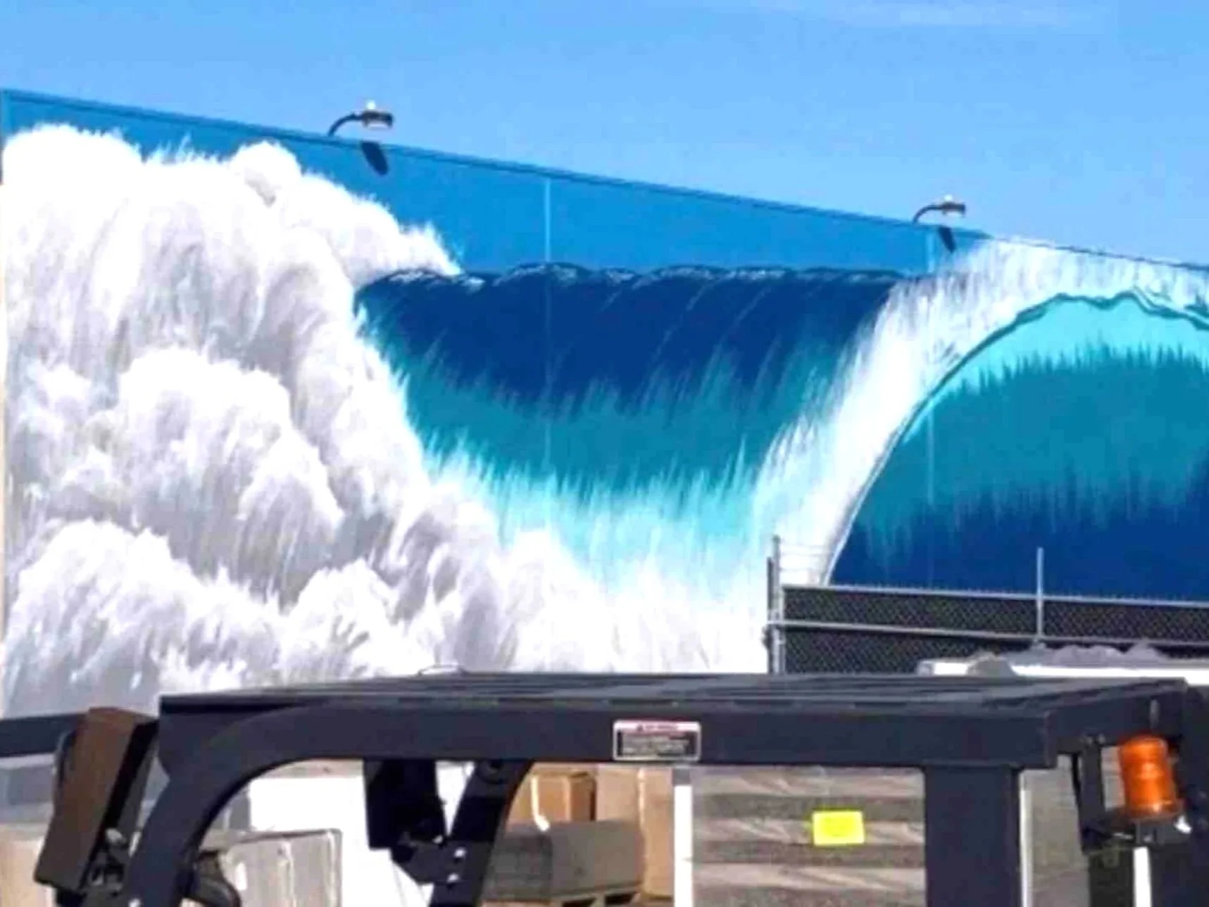 A large mural of a wave is painted on a building wall, with water cascading down the edge of the wave and creating white foam, against a clear blue sky.