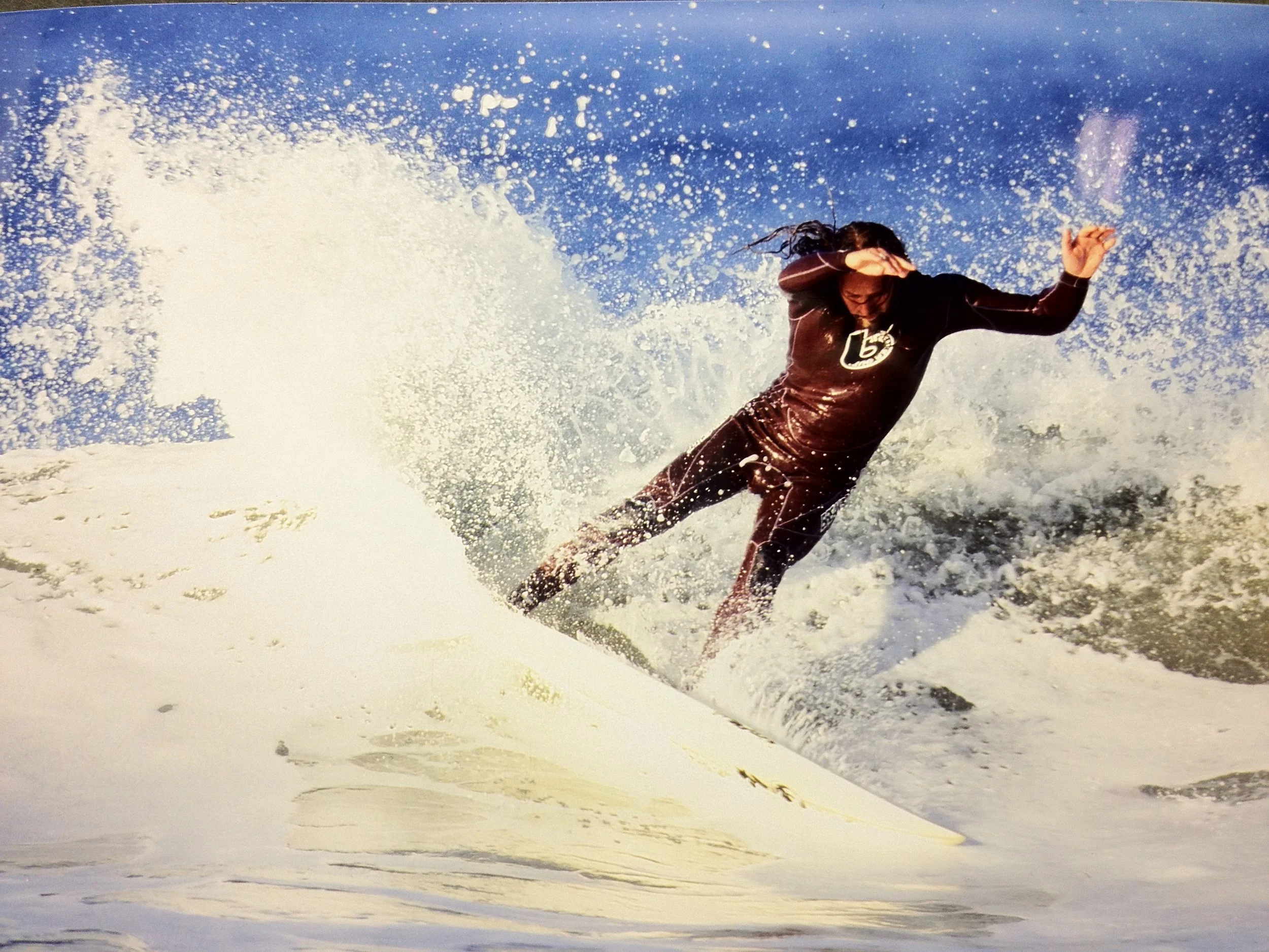 John Glomb surfing on a wave in the ocean, wearing a dark wetsuit, with spray of water around him.
