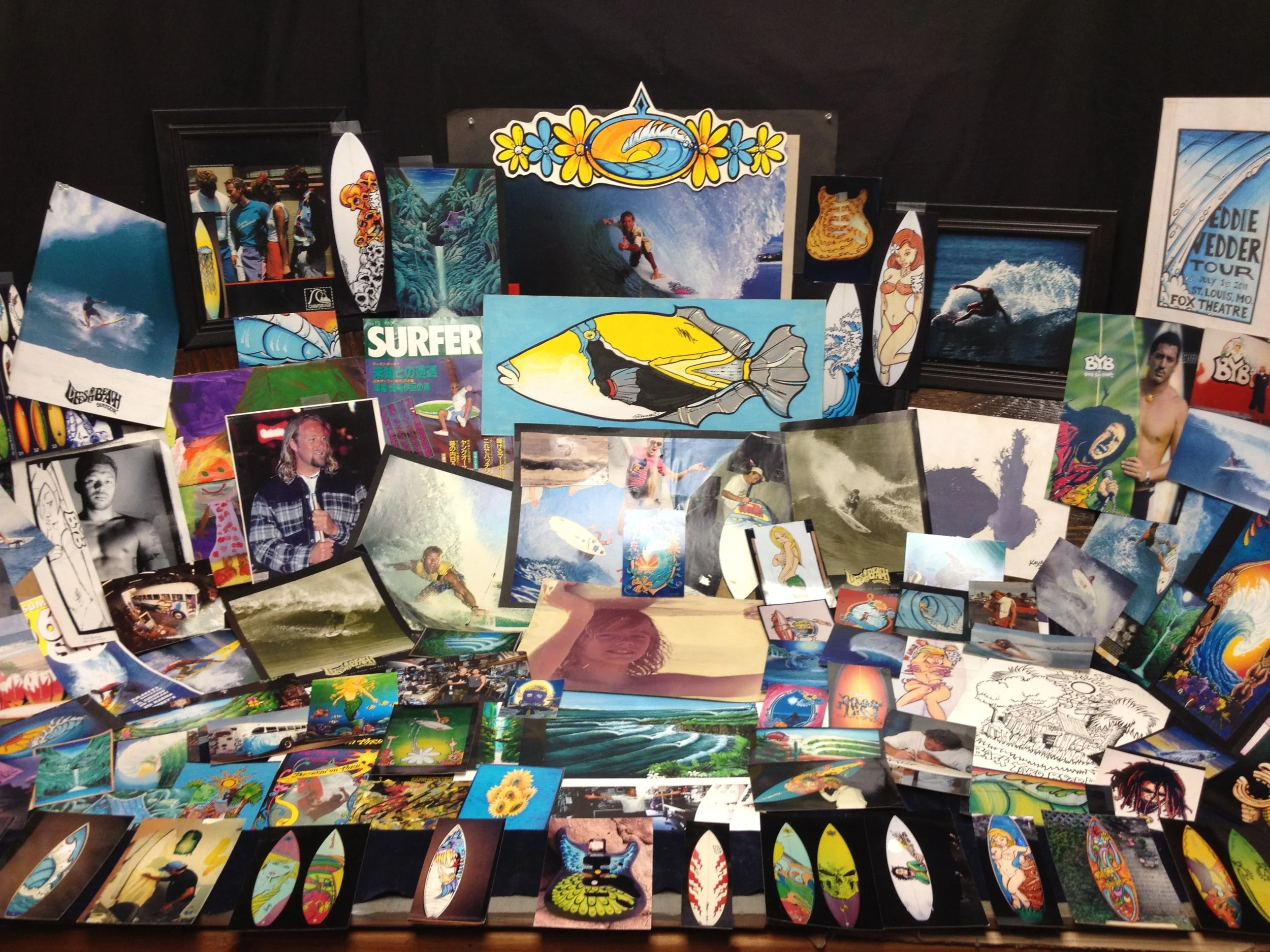 A collection of surfing-themed photographs, paintings, and illustrations displayed on a table and pinned on a wall, featuring surfers, waves, surfboards, and related artwork.