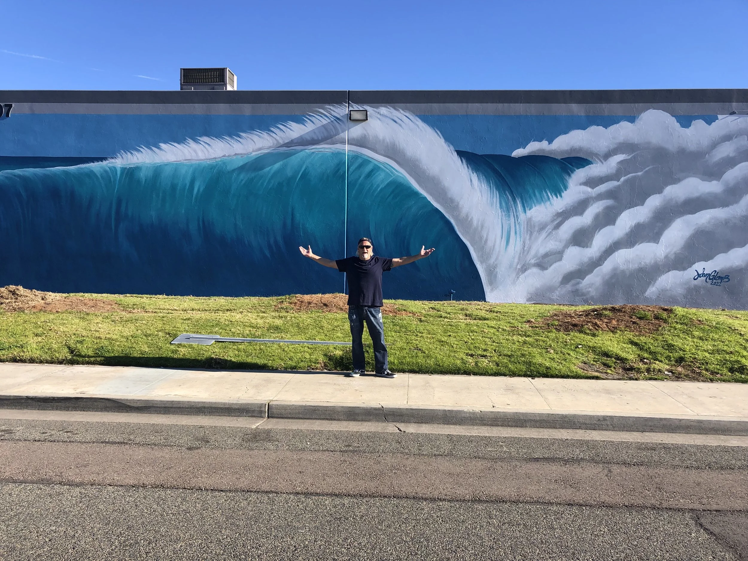 John Glomb standing with arms outstretched in front of a large mural of a giant ocean wave on a building wall during daytime.