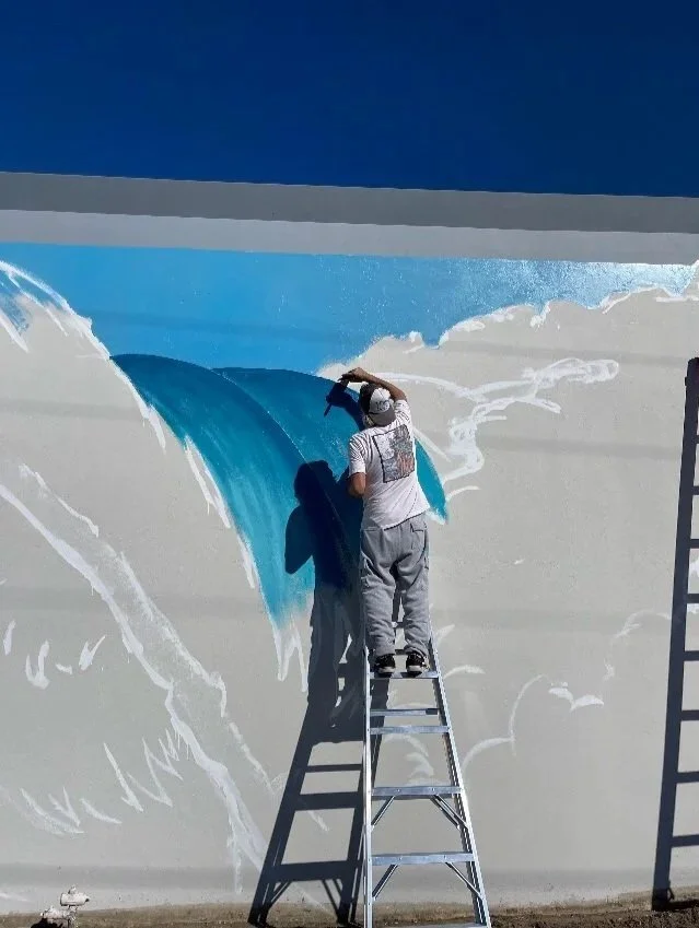 John Glomb painting a mural of a wave on a wall, standing on a ladder