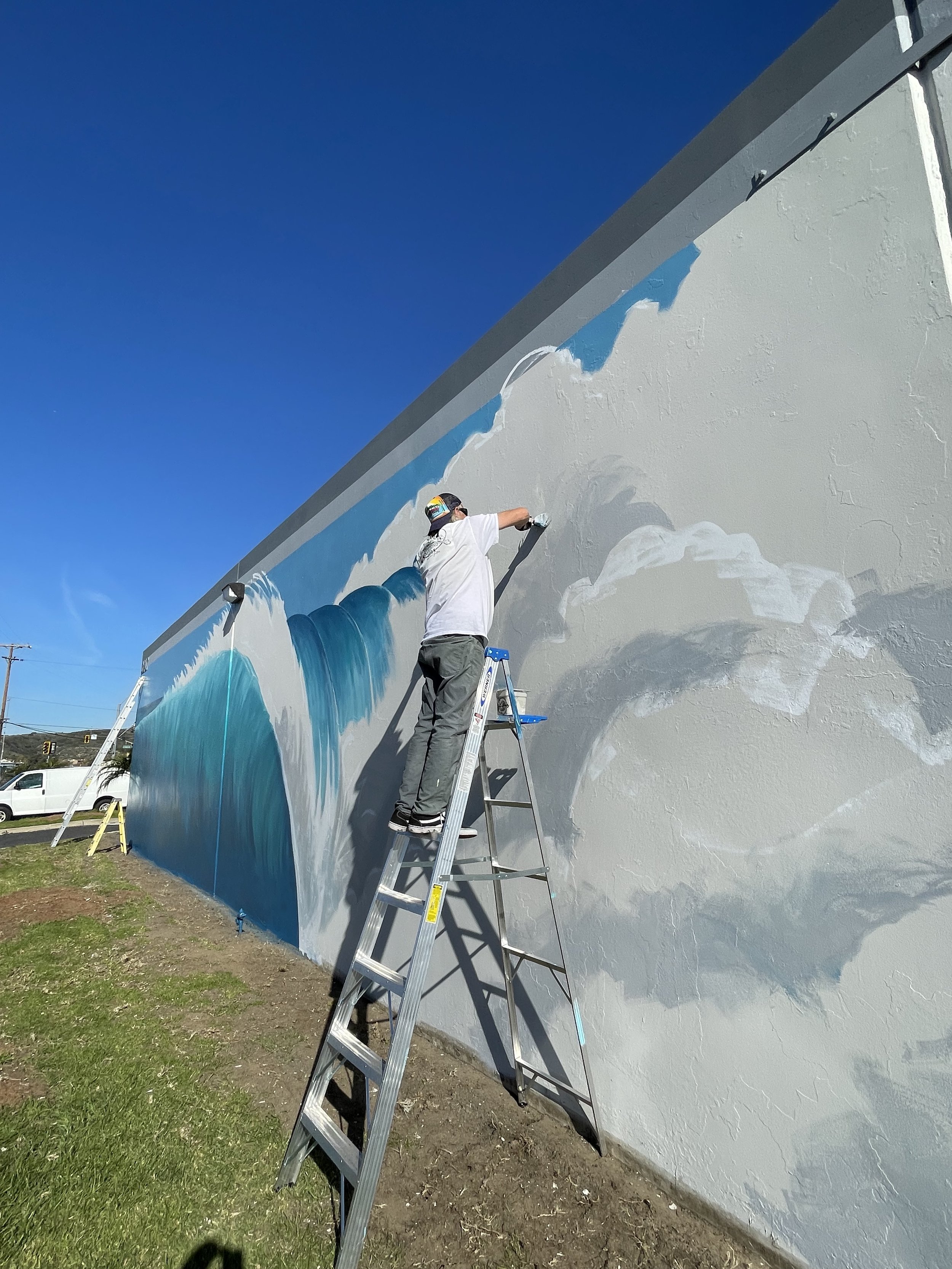 John Glomb painting a mural of ocean waves on a large outdoor wall, standing on a ladder with a blue sky background.
