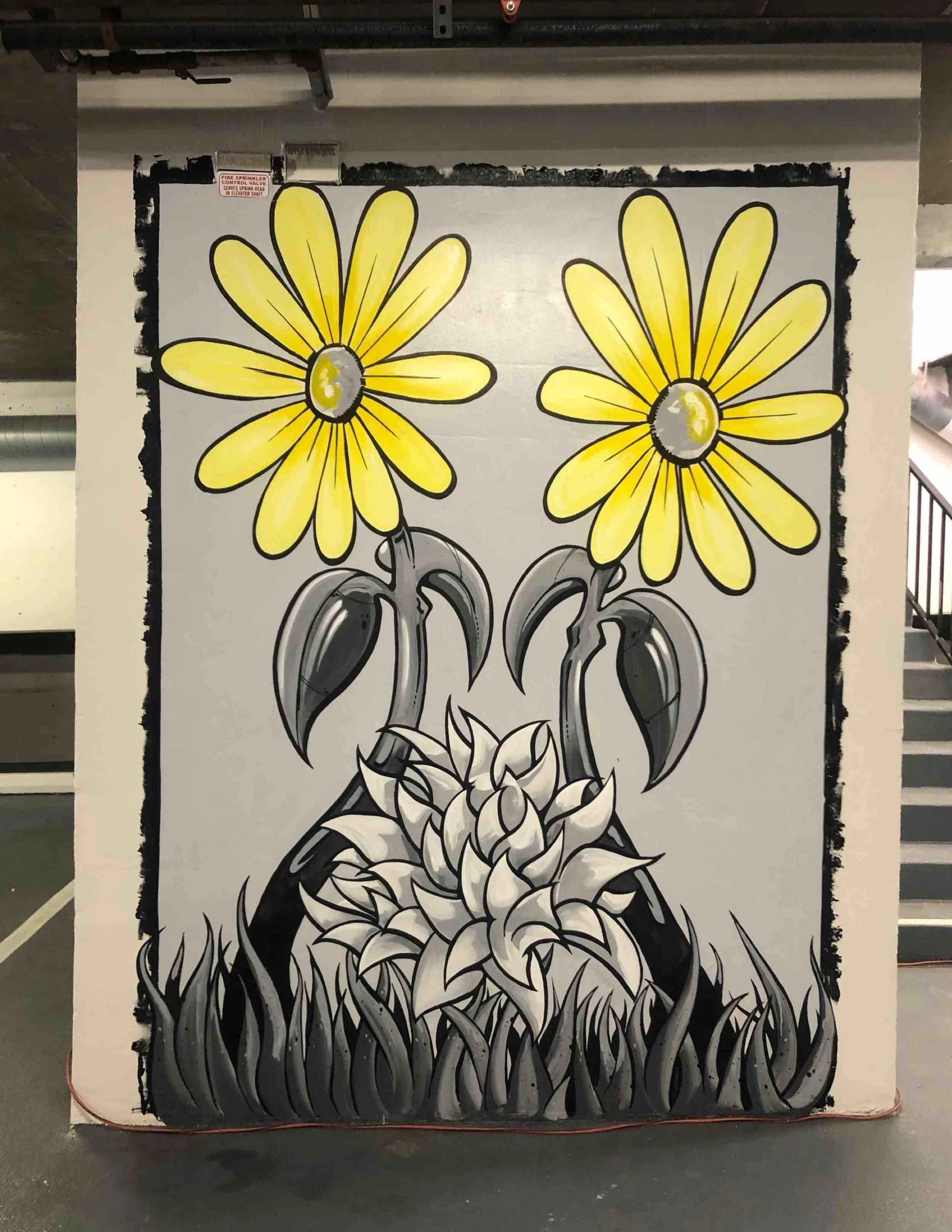 Street art mural of two black and white hand holding a flower with large yellow daisy-like flowers with yellow centers and black outlines, set against a gray background.