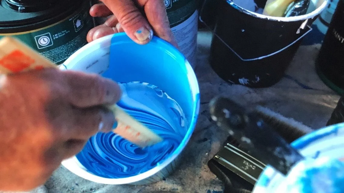 Close-up of a person mixing blue paint in a white bowl with a wooden stick, surrounded by various paint cans and containers on a work surface.