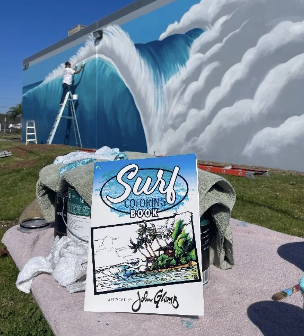 A mural artist is painting a large, realistic wave on a building wall. Art supplies and a 'Surf Coloring Book' are on a table in the foreground.