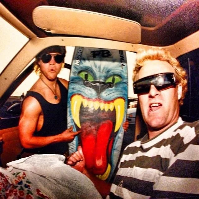 John Glomb and Brad Gerlach wearing sunglasses in a car, holding a large skateboard with a fierce, colorful cat face design and open mouth. John Glomb has blond hair and wears a striped shirt, and Brad has dark hair, a black tank top, and necklaces.