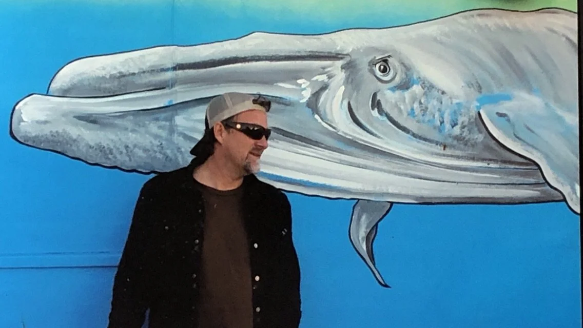 John Glomb wearing sunglasses, a cap backwards, and a dark jacket standing in front of a large mural of a whale painted on a blue background.