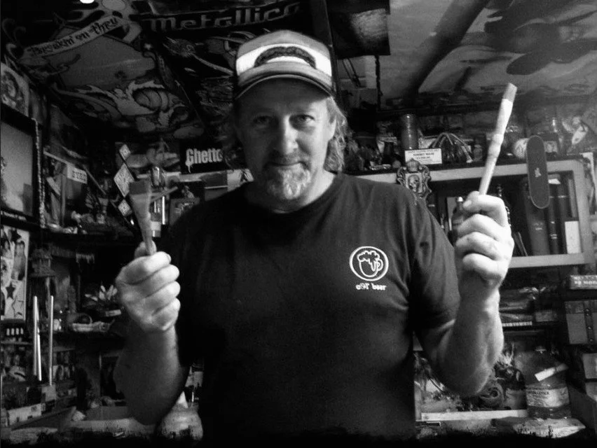 John Glomb with a beard and long hair wearing a baseball cap and a T-shirt with a logo, holding a paintbrush in each hand, standing in a cluttered room filled with posters, books, and various objects.