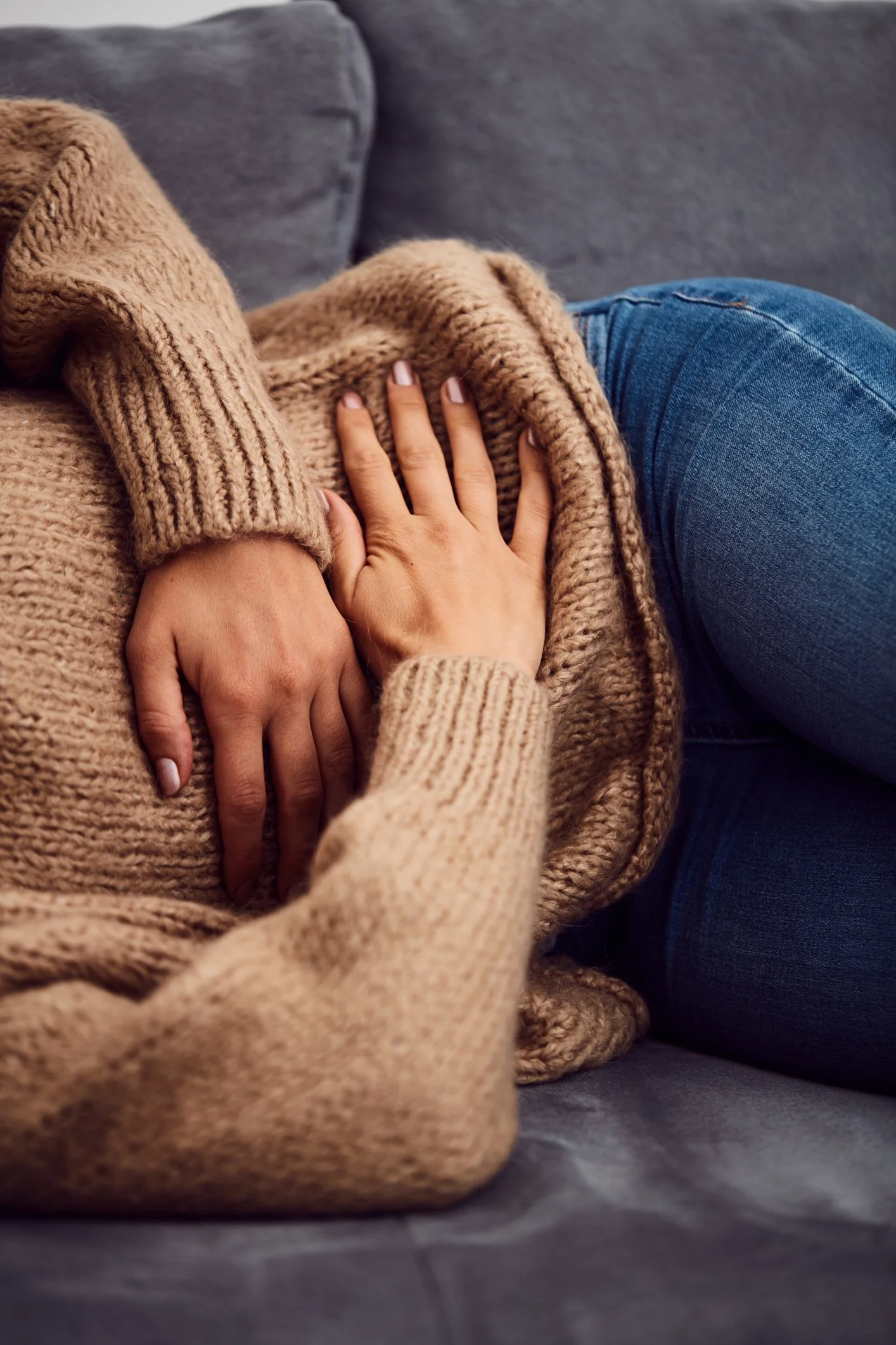 Person wearing a beige sweater, lying on a gray couch, with their hand on their stomach.