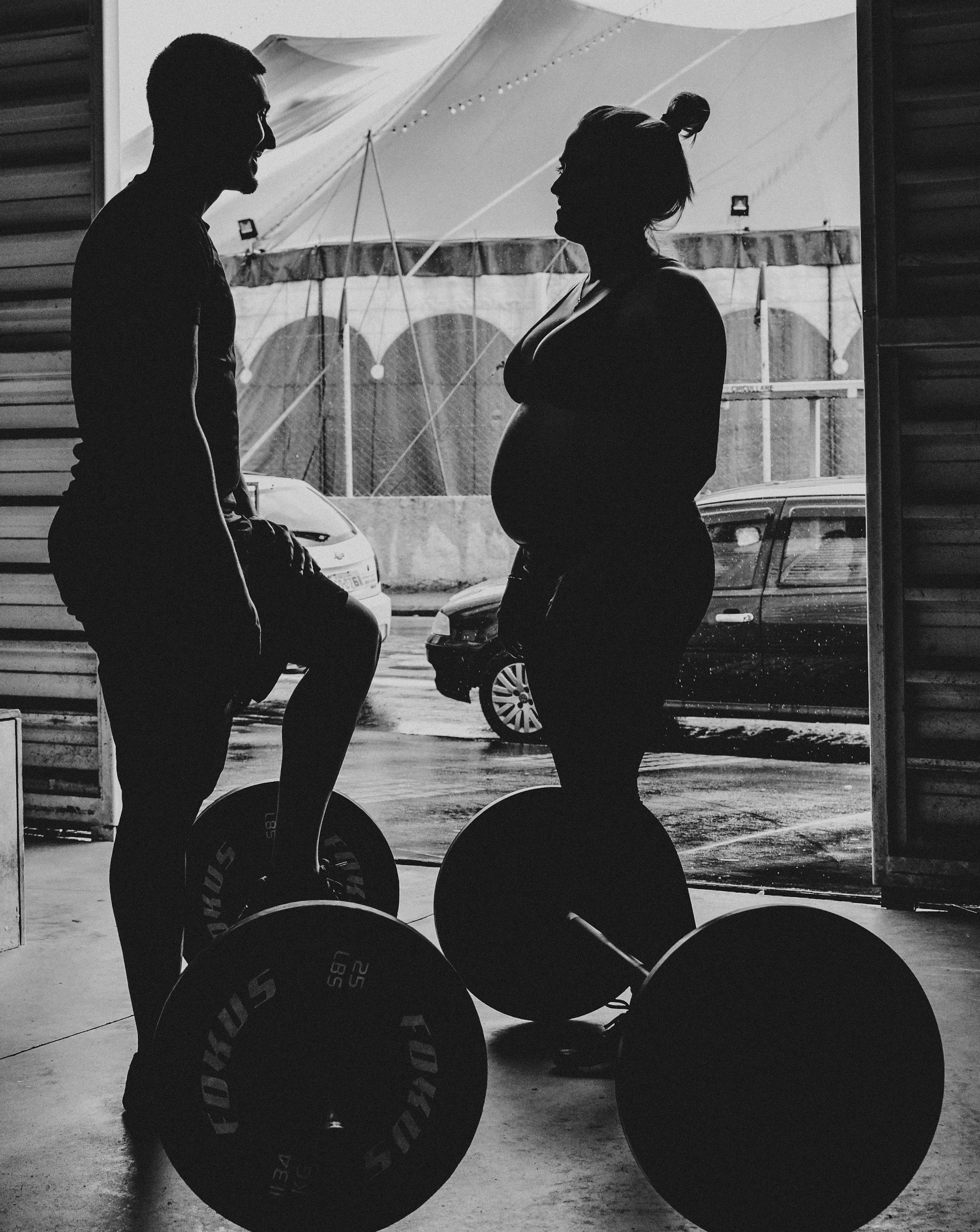 Silhouettes of a man and a pregnant woman talking in a gym parking lot, with weightlifting equipment and cars visible outside.