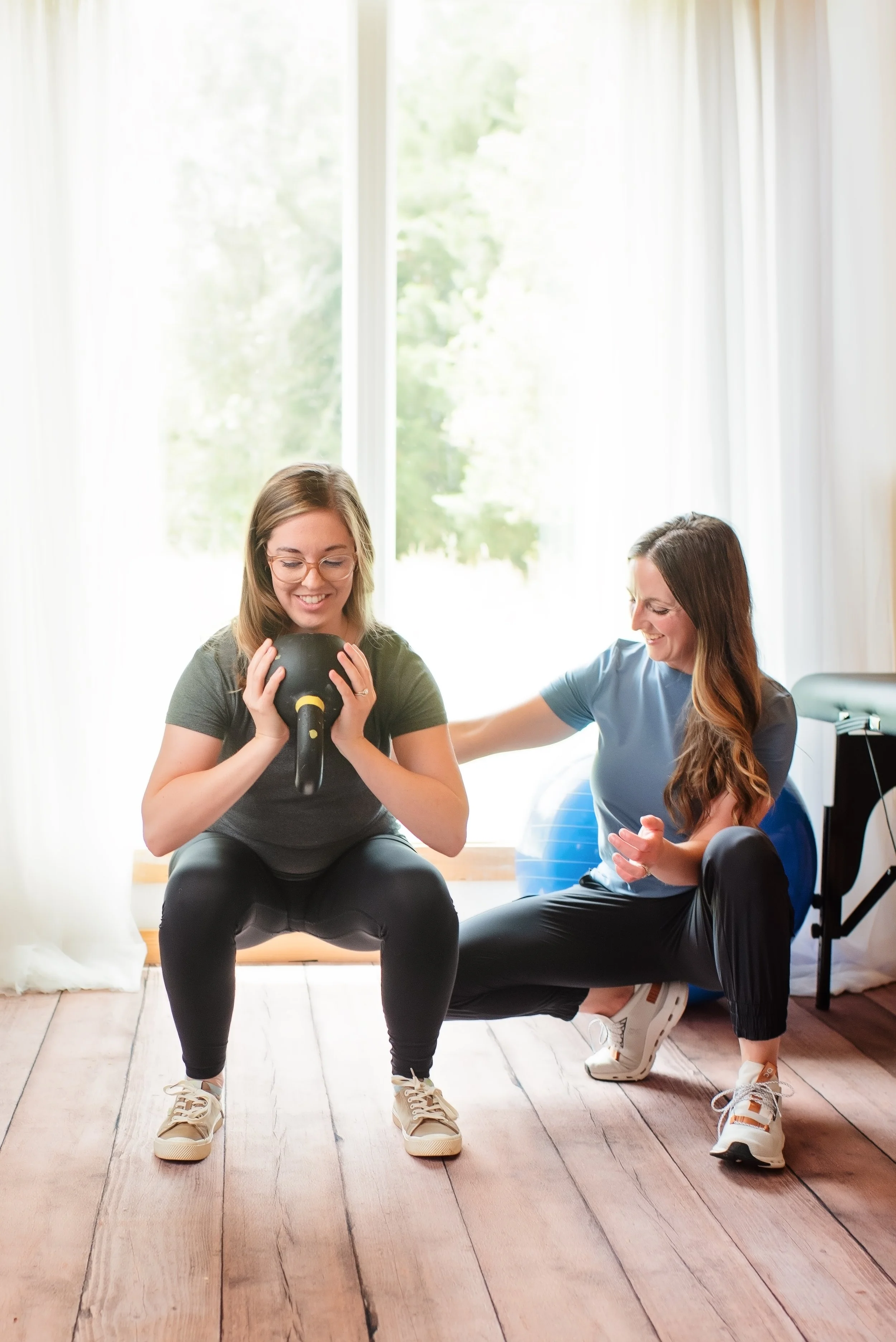 Two women exercising indoors with one using a kettlebell and the other as a trainer supporting her, with a large window and fitness equipment in the background.