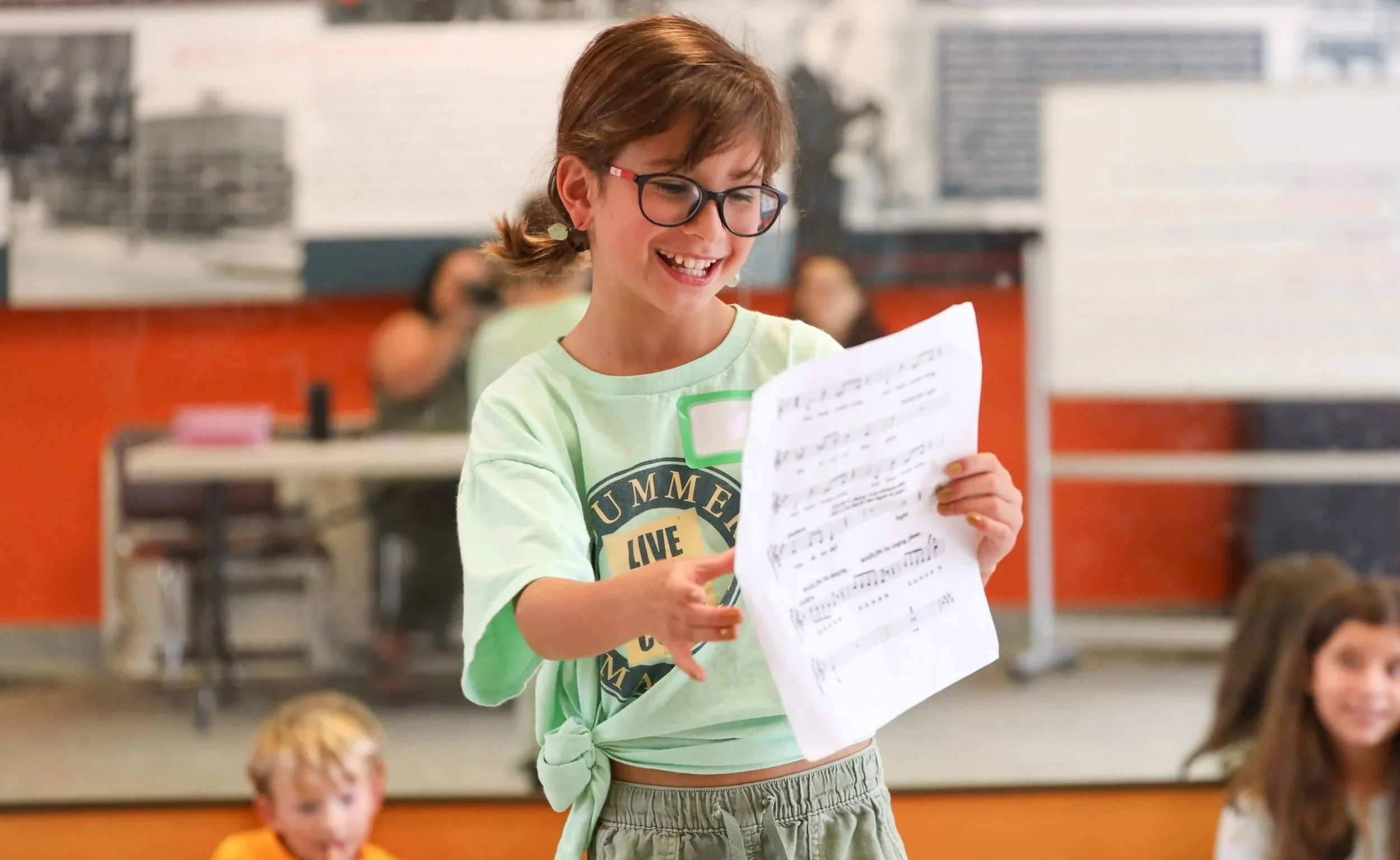A young girl with glasses and a green t-shirt reading music notes on paper in a classroom or auditorium setting, with other children seated around her.