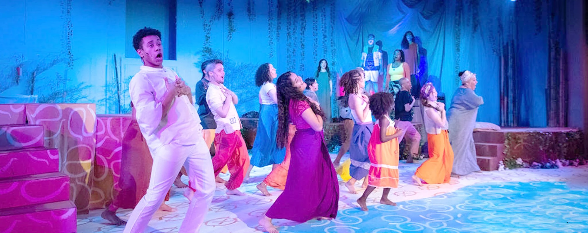 A diverse group of people performing on stage in colorful costumes, with a blue backdrop and stage props.