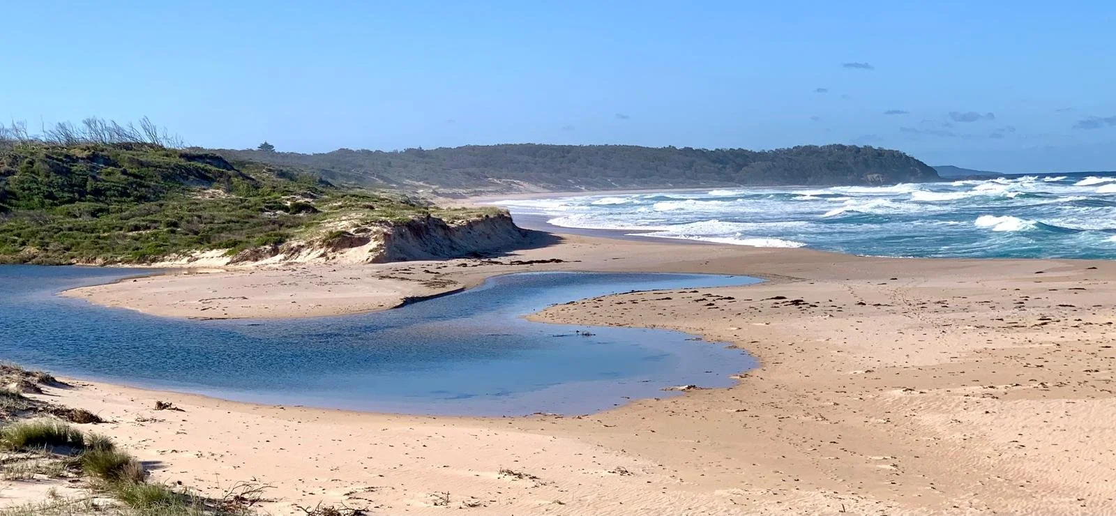 An image of a beach with a river running down past a sand dune towards the sea.
