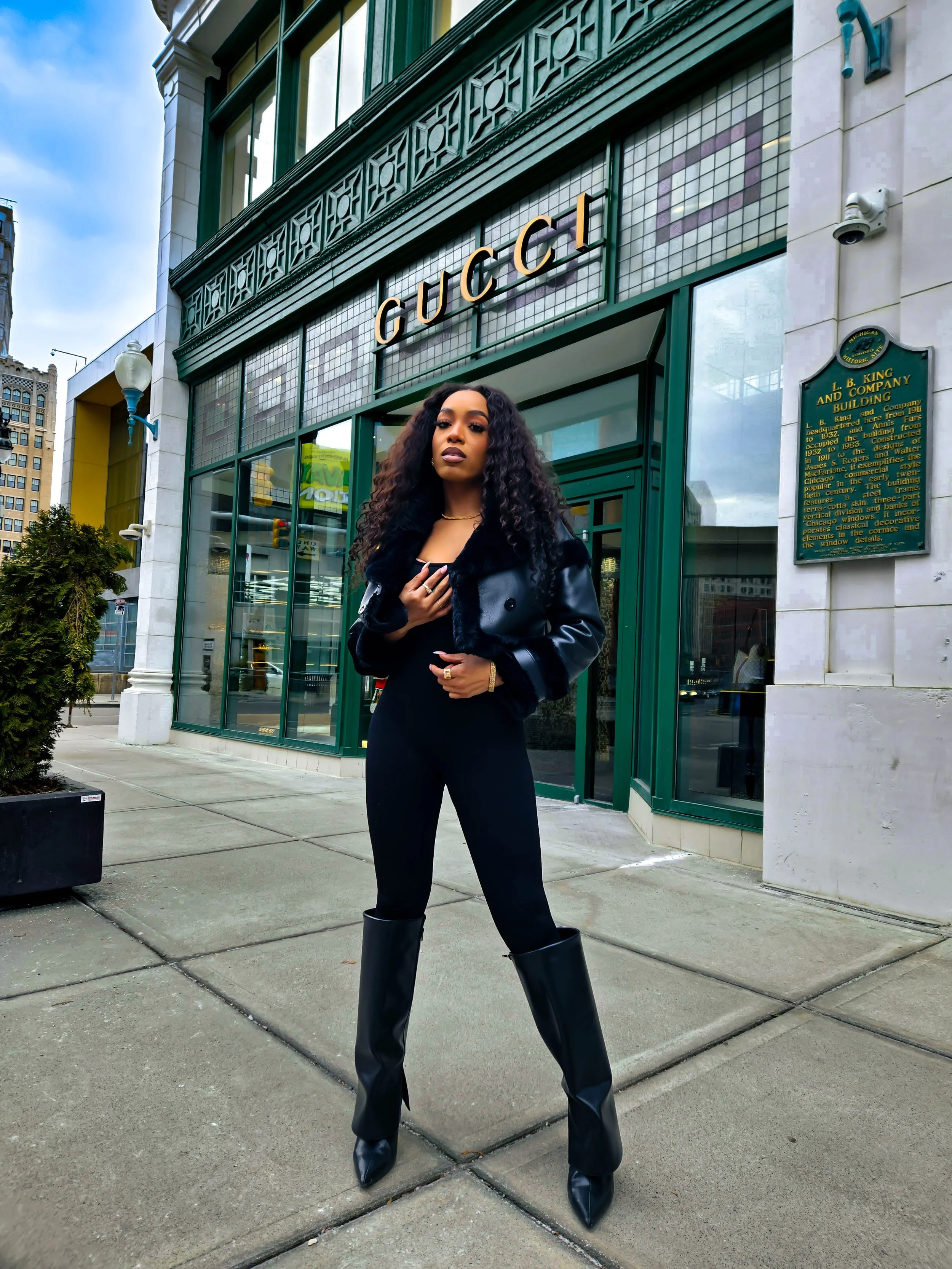 A woman with curly hair standing on a city sidewalk in front of a Gucci store, wearing a black jacket, black leggings, and tall black boots.