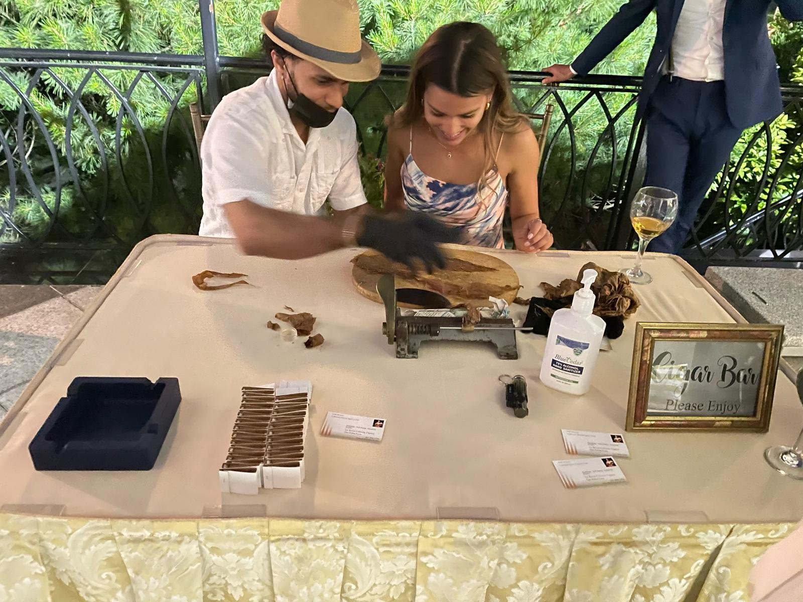 A man and woman sit at a table in a cigar bar, watching a cigar cutter in operation. The man wears a beige hat and a black mask, while the woman is smiling. The table has a sign that says "Cigar Bar Please Enjoy" and a glass of white wine.