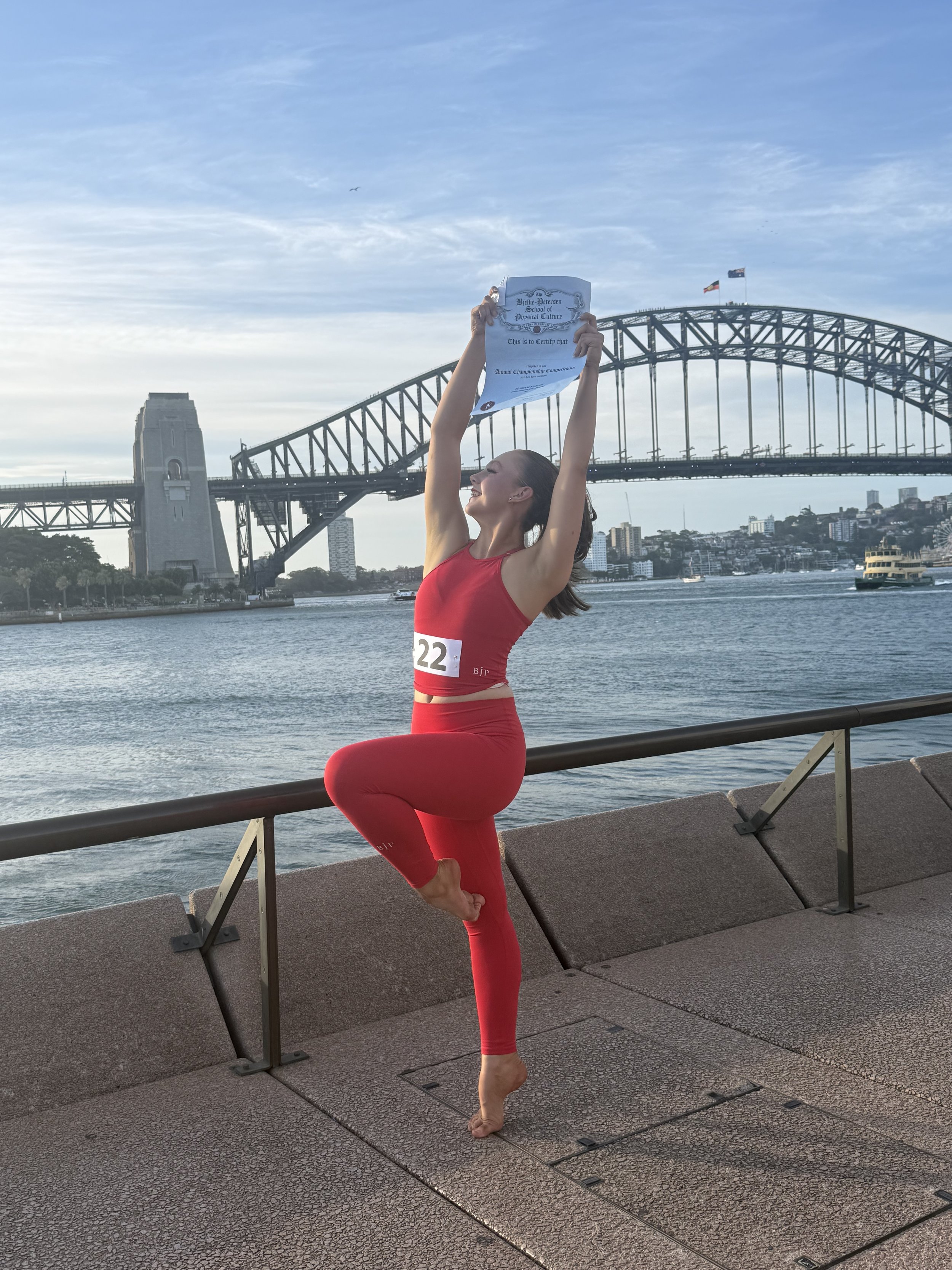 A woman in a red athletic outfit practicing yoga on a waterfront promenade with the Sydney Harbour Bridge in the background, holding a certificate overhead and balancing on one leg.