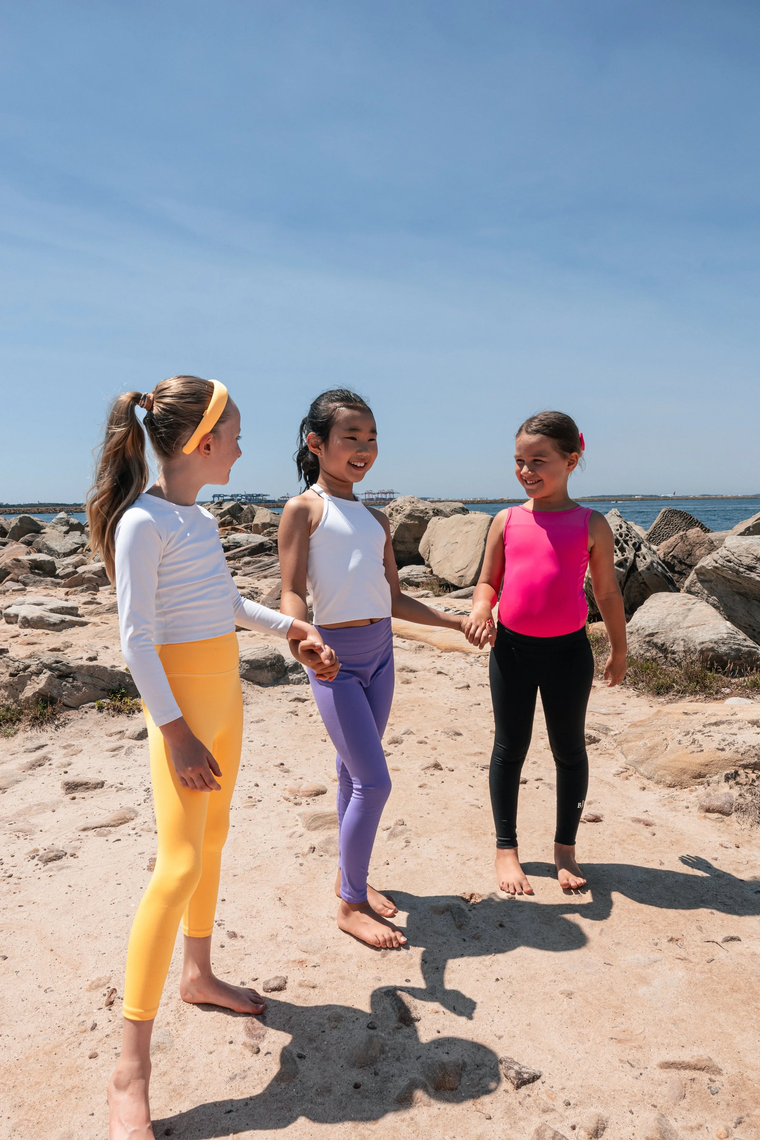Young girl in a white athletic outfit performing a dance or gymnastic move on a wooden floor, with her arms extended and one leg bent, in an indoor setting.