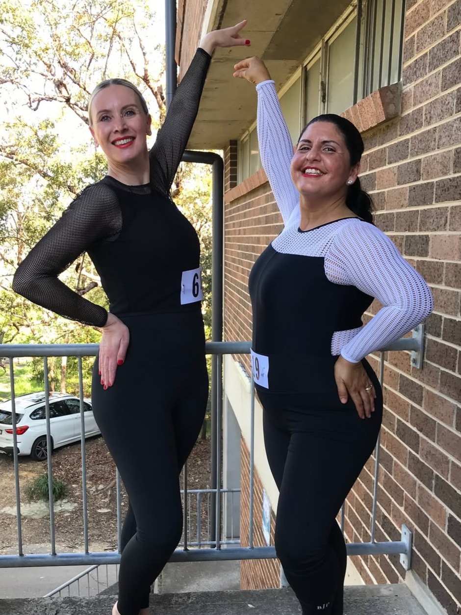 Two women smiling and posing together in a gymnasium, wearing workout clothes and competition bibs, with numbers 72 and 10.