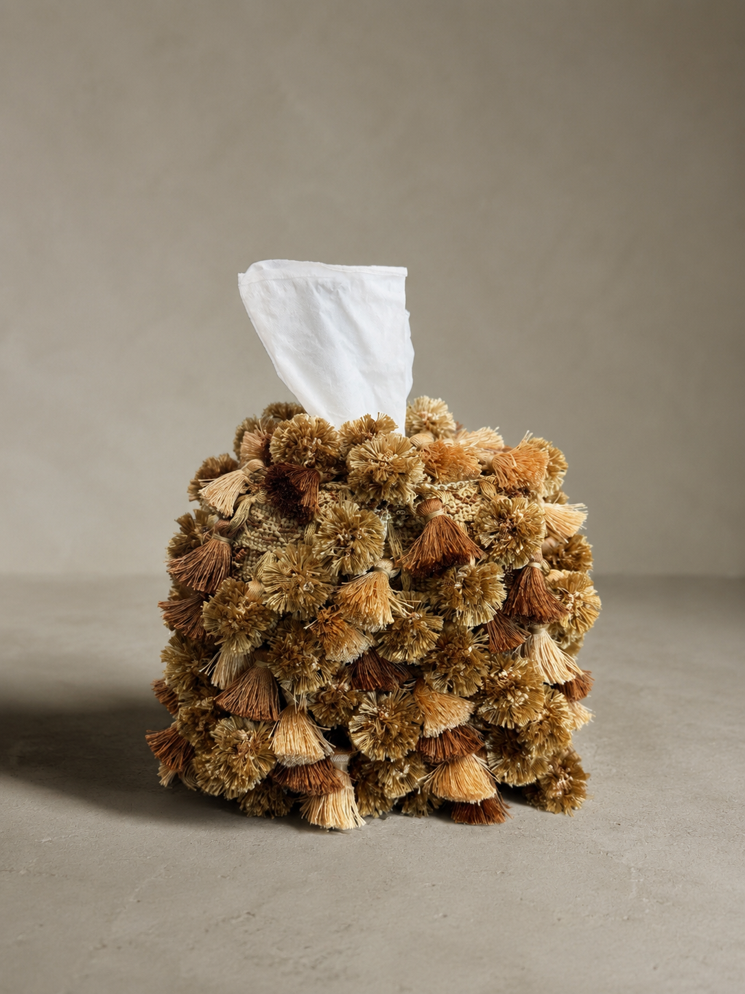 Close-up of a handcrafted tissue box cover with dense tassel fringe in varying shades of soft green, golden tan, and rich brown.