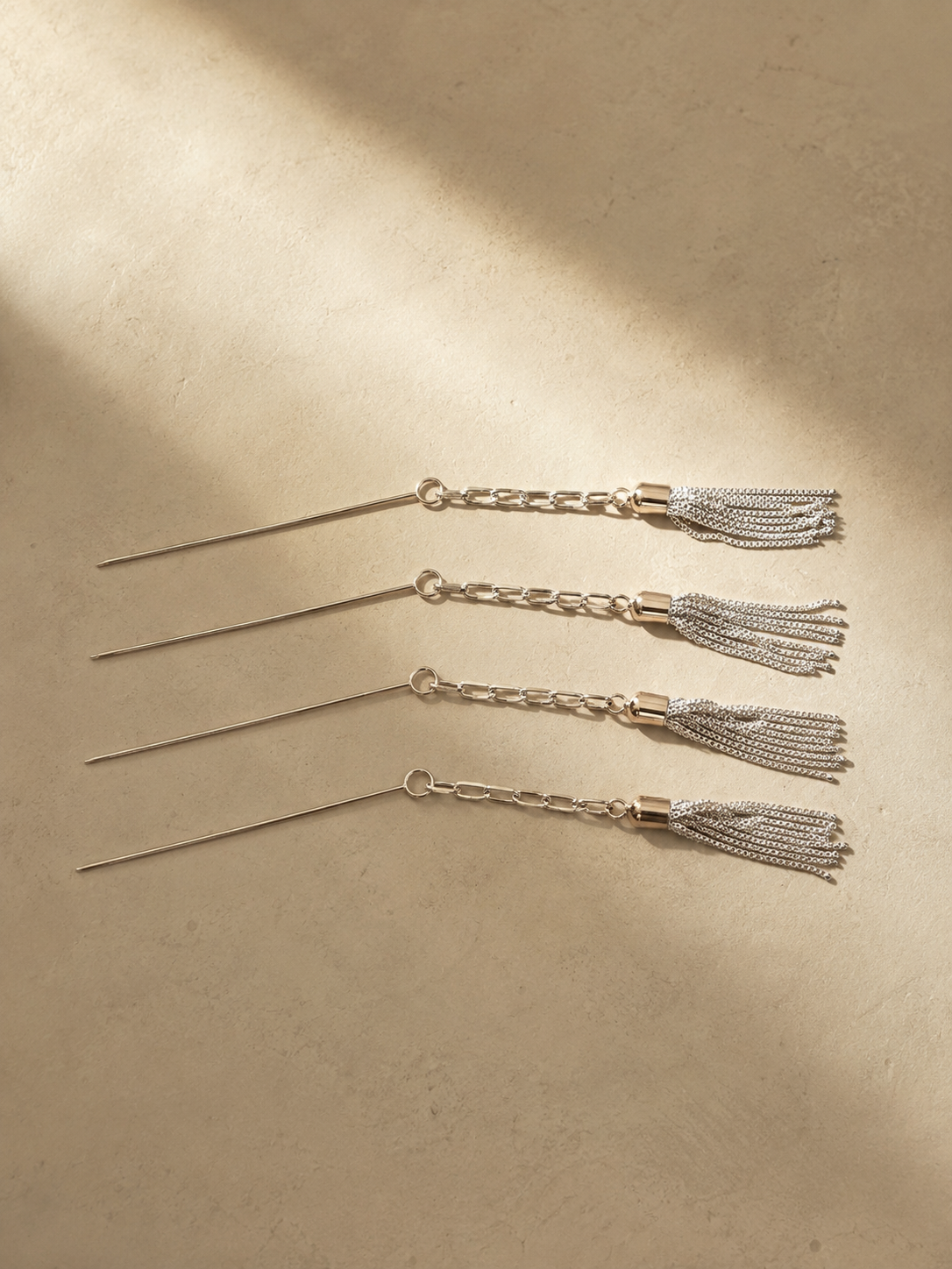 Set of four stainless steel Tassel Cocktail Picks with silver-plated brass tassels displayed in a stainless steel holder on a warm stone surface with soft editorial lighting.