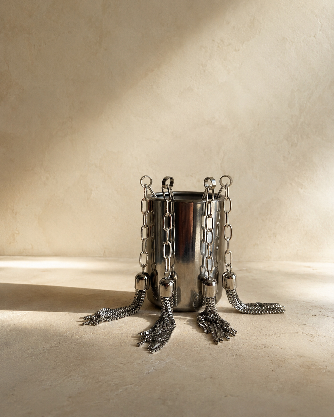 Set of four stainless steel Tassel Cocktail Picks with silver-plated brass tassels displayed in a stainless steel holder on a warm stone surface with soft editorial lighting.