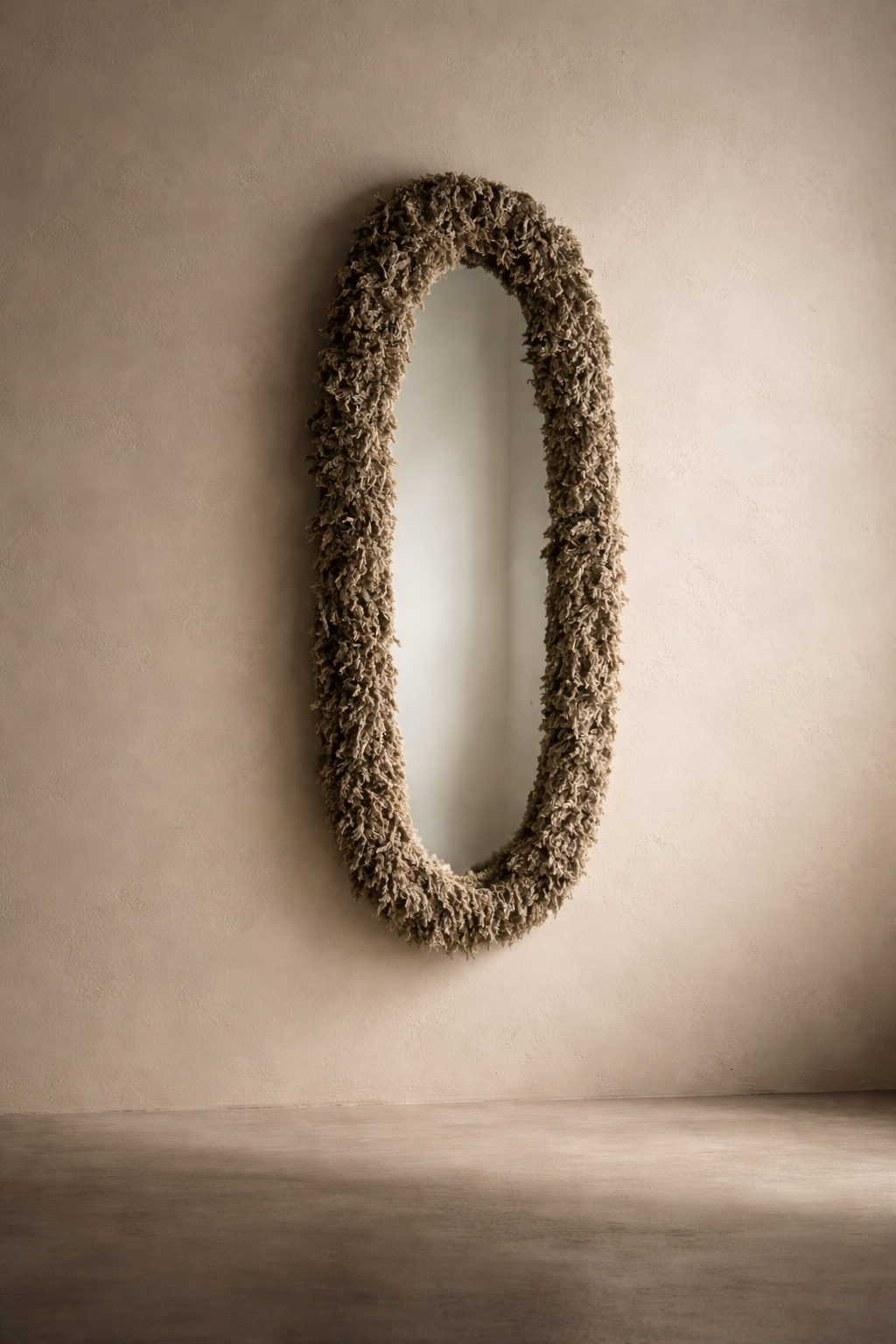 Elongated oval wall mirror wrapped in soft moss shag fringe, shown against a warm neutral plaster wall in soft natural light.