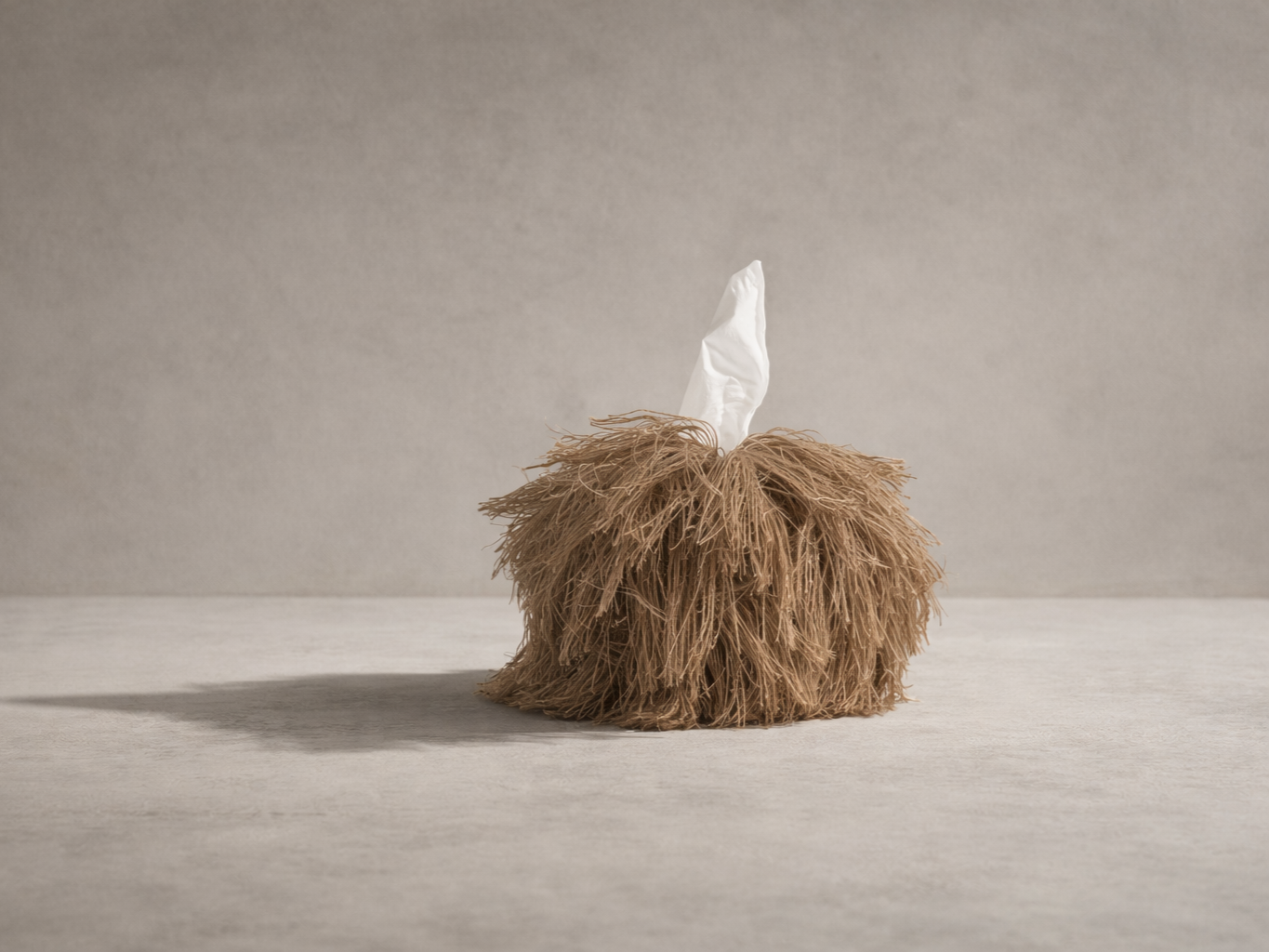 Rooted jute fringe tissue box cover by Amber Renee Hansen on warm neutral plaster floor with directional lighting.