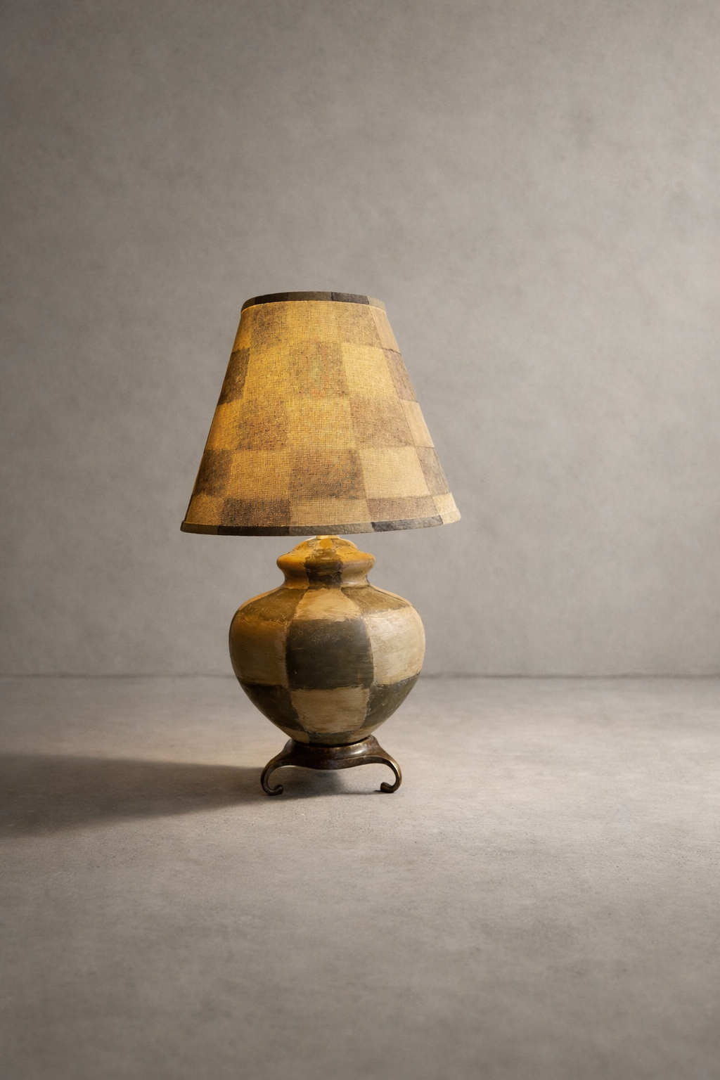 Neutral checkered shade lamp with tassel fringe and a column base on a brass scroll pedestal against a plaster wall.