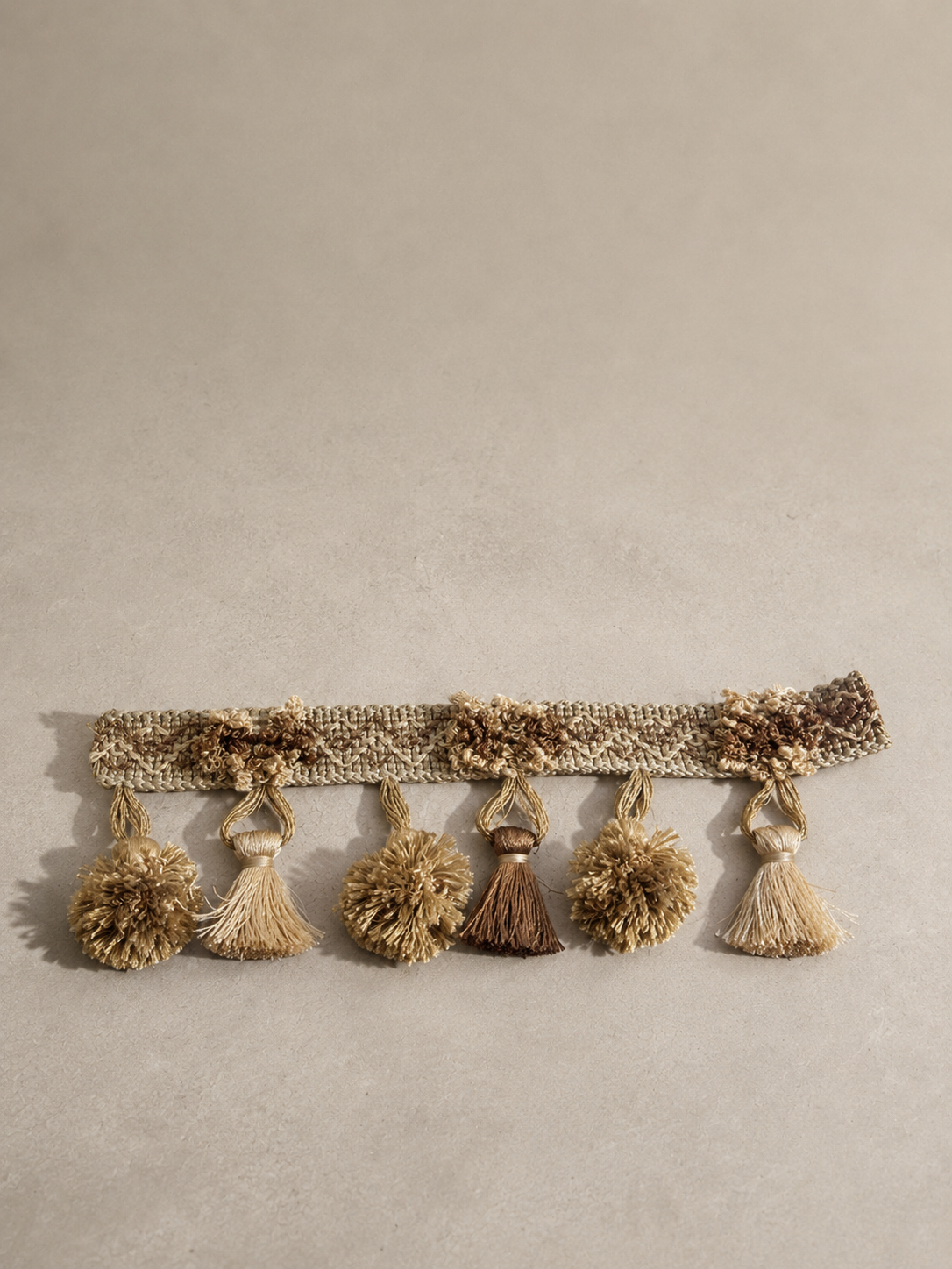 The Gathered Earth Tassel Tissue Box Cover sits on a neutral studio surface, showcasing its olive, tan, and brown fringe tassels with a sculptural, textured appearance.