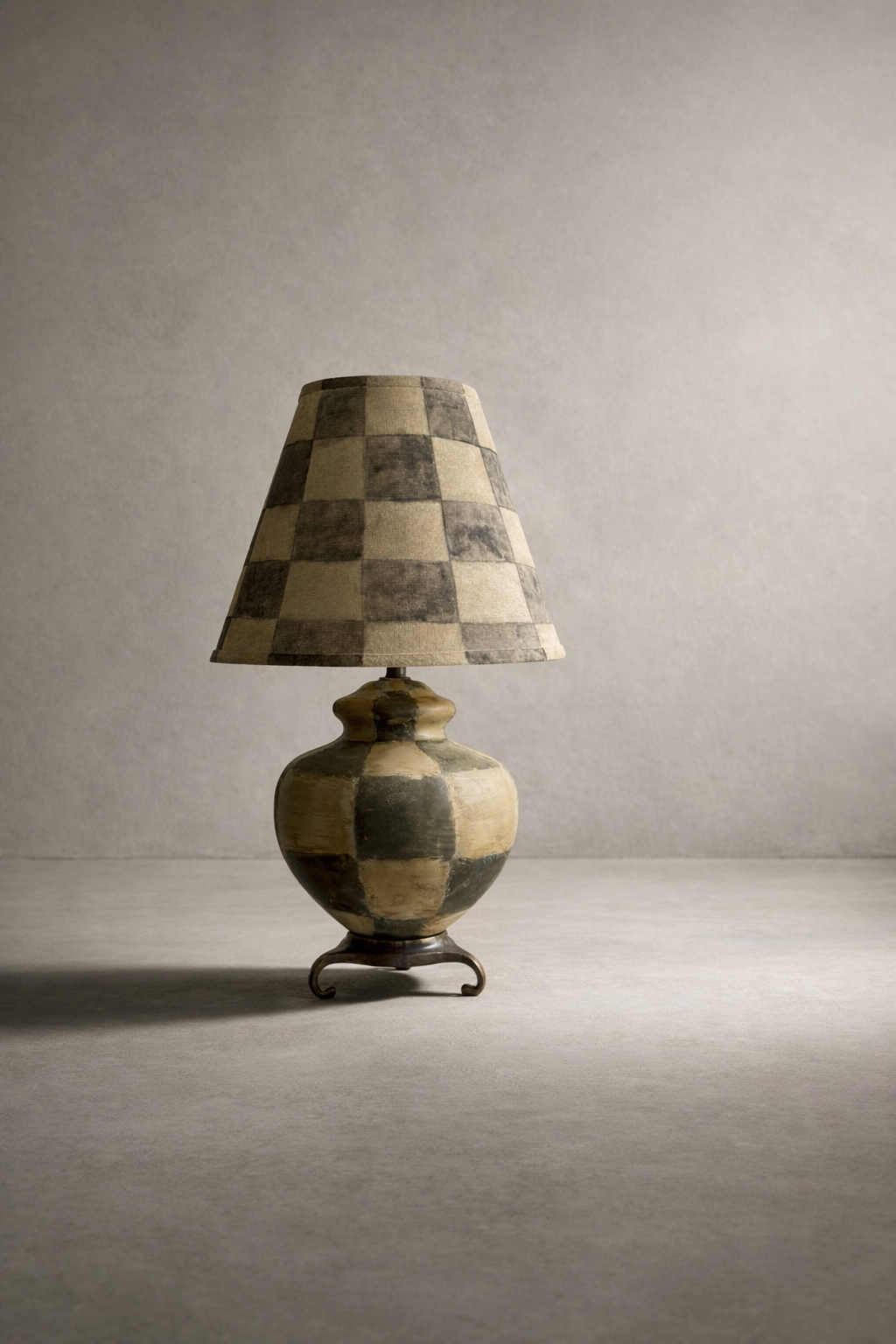 Neutral checkered shade lamp with tassel fringe and a column base on a brass scroll pedestal against a plaster wall.