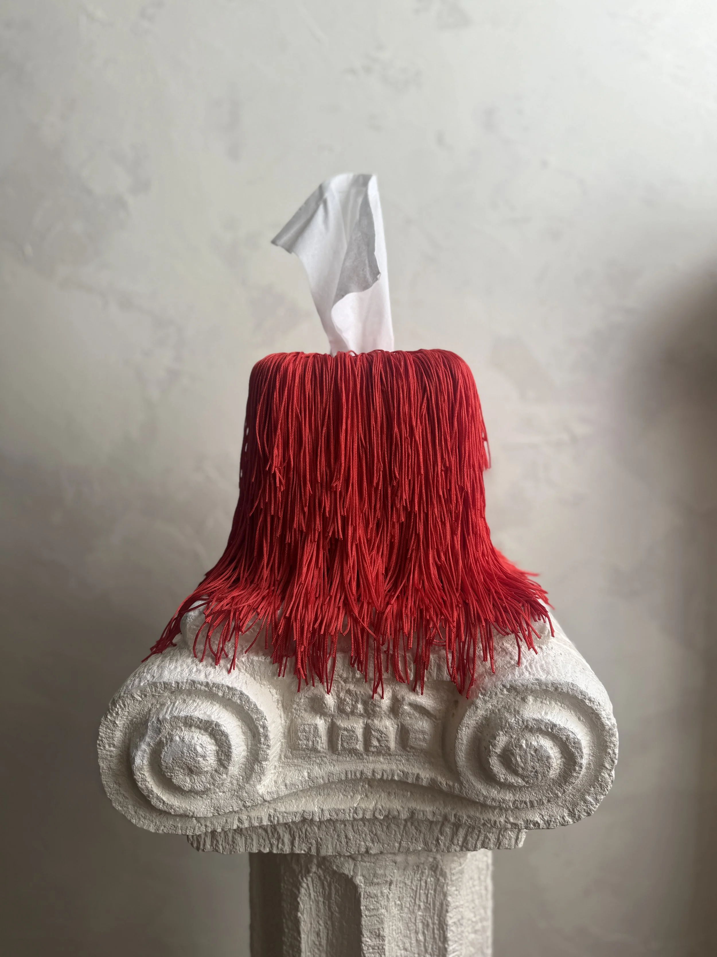 Fringe tissue box cover in warm vermillion red with soft draped fringe pooling at the base, styled in a minimal neutral interior.