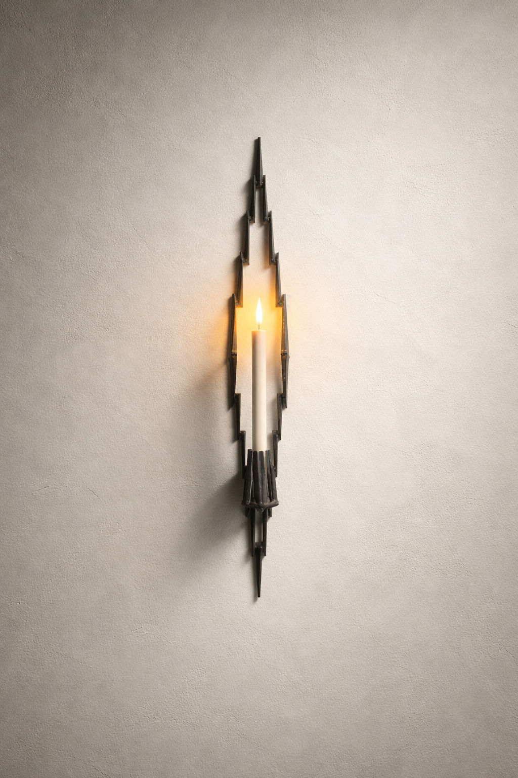 Hand-welded steel vertical wall candle sconce holding a lit taper candle, casting sculptural shadows on a plaster wall