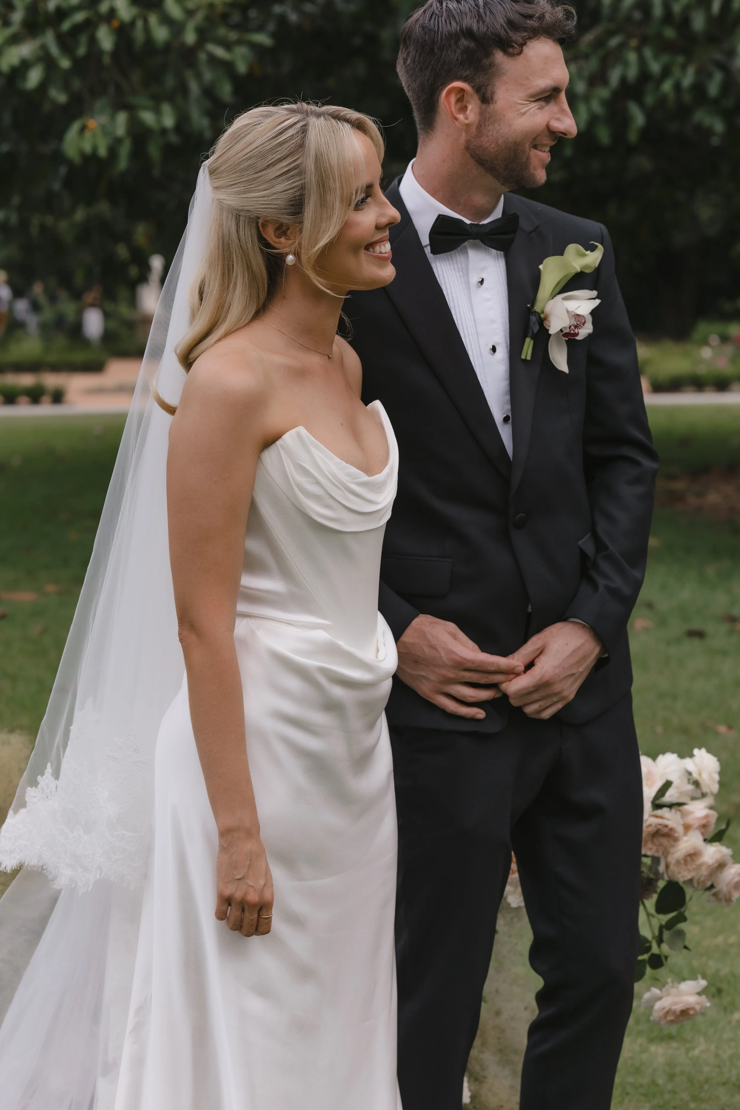 A bride and groom standing side by side outdoors during a wedding ceremony, smiling. The bride is wearing a strapless white wedding dress with a veil, and the groom is in a black tuxedo with a bow tie and a white flower boutonniere. Green trees and a grassy area are visible in the background.