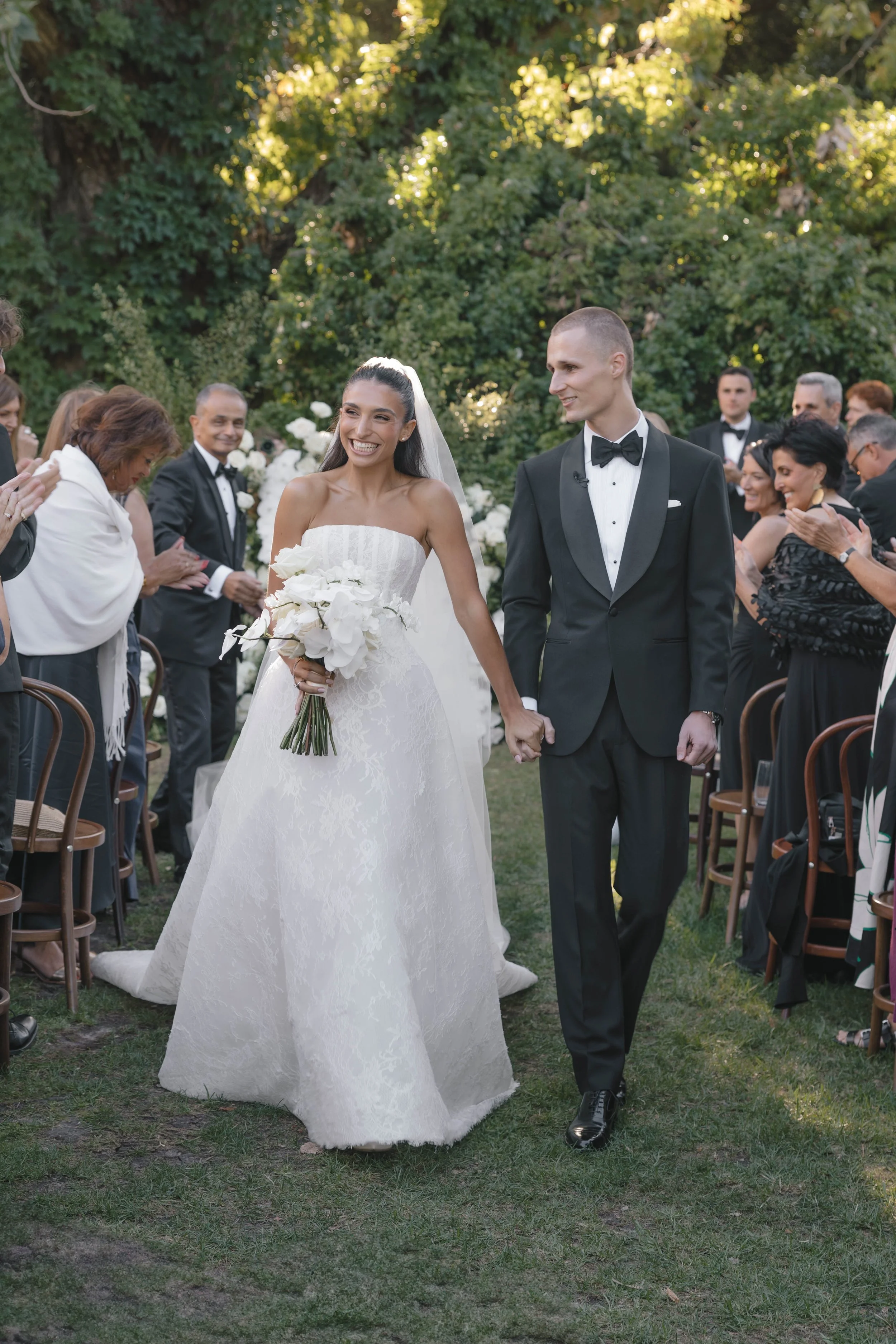 A bride and groom walking hand in hand down an outdoor aisle, smiling, surrounded by wedding guests.