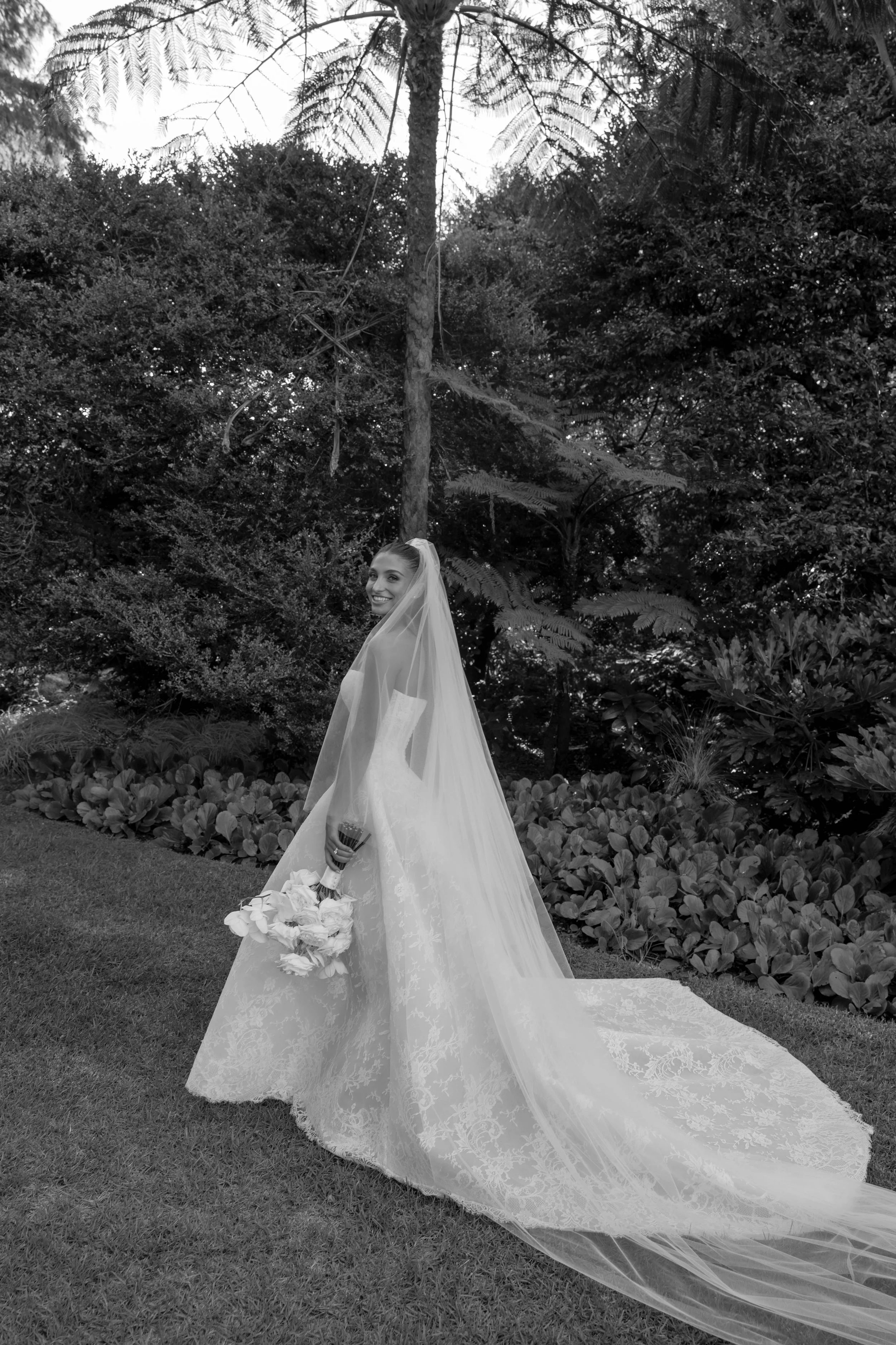 A smiling bride in a wedding dress with a long lace train and veil holding a bouquet of flowers standing on a lawn with lush bushes and trees in the background.