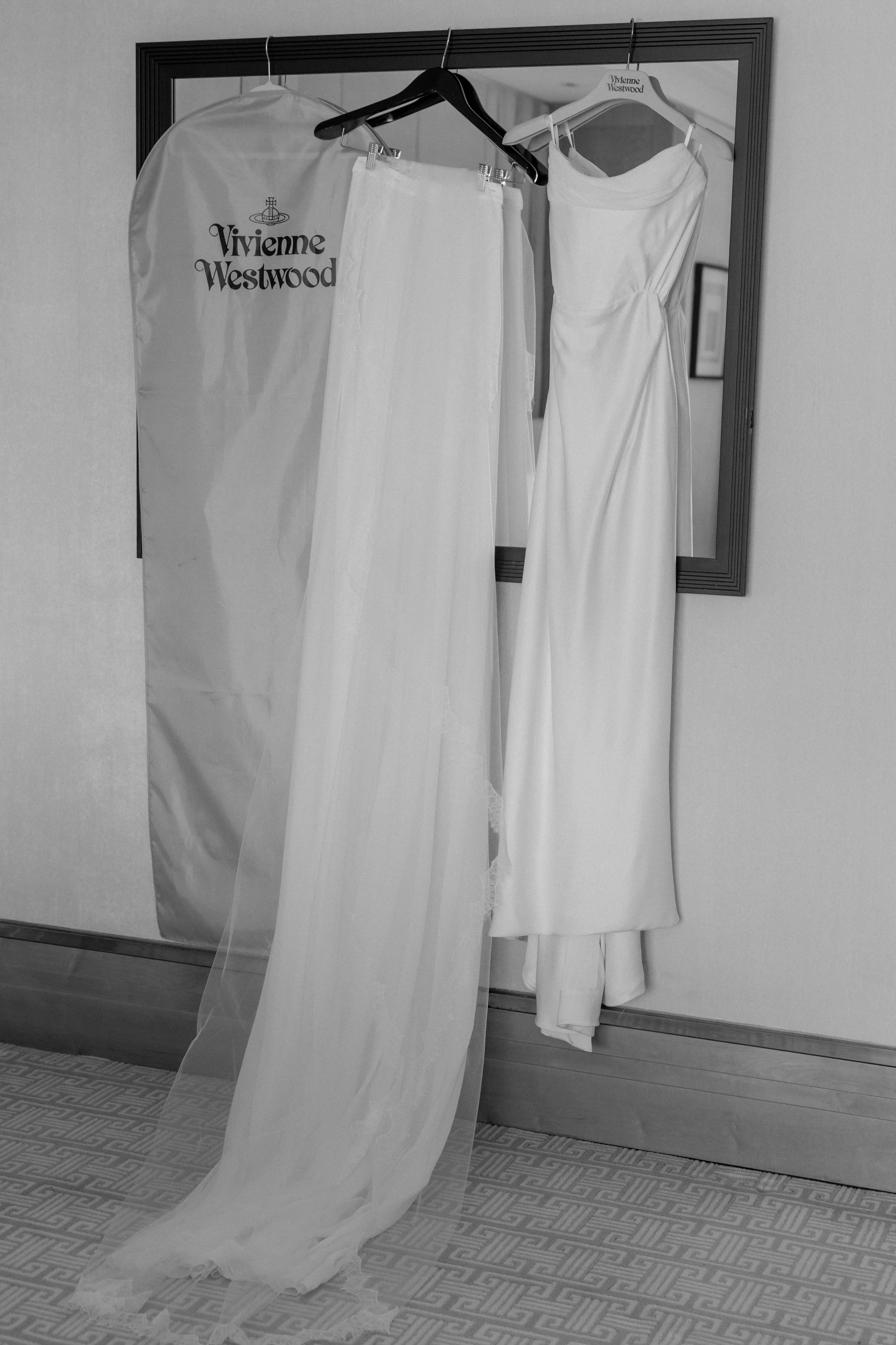 White wedding dress and veil hanging on hangers in front of a mirror with a Vivienne Westwood garment bag nearby.