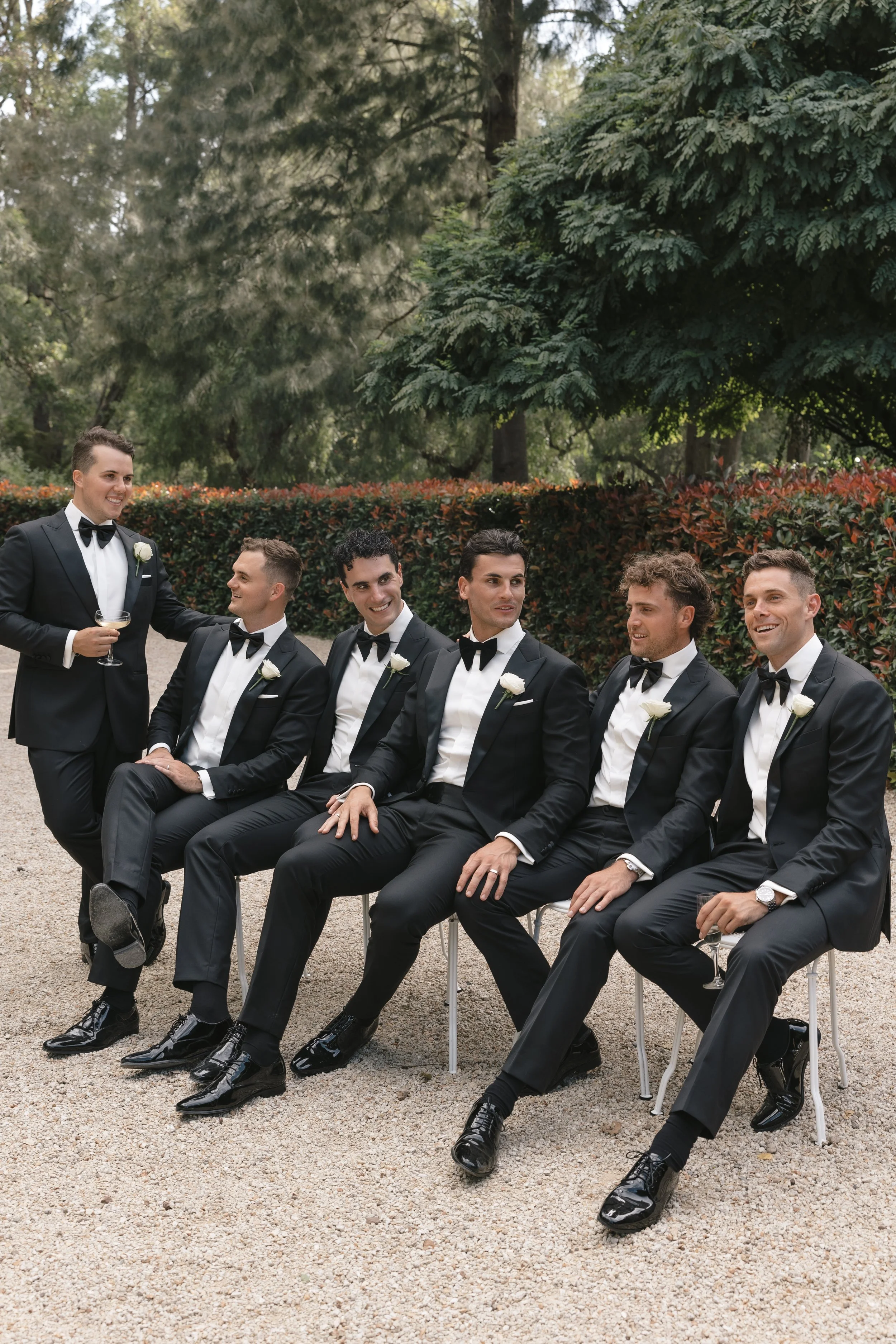 Group of six men in black tuxedos with white shirts and black bow ties, sitting and standing outdoors on gravel, surrounded by green trees and bushes, celebrating at a formal event.