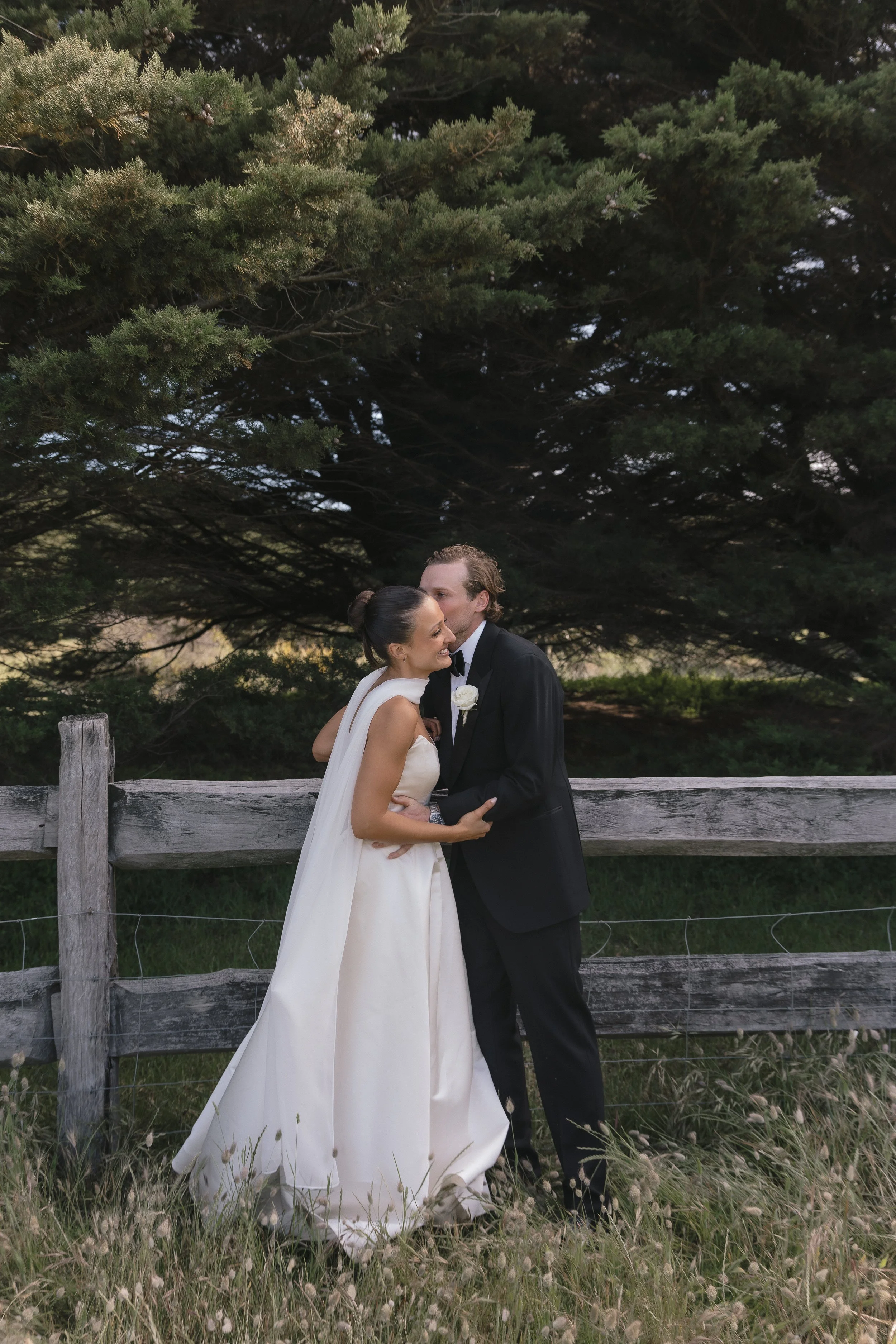 A bride and groom sharing a kiss outdoors in front of a wooden fence and large trees, dressed in wedding attire.