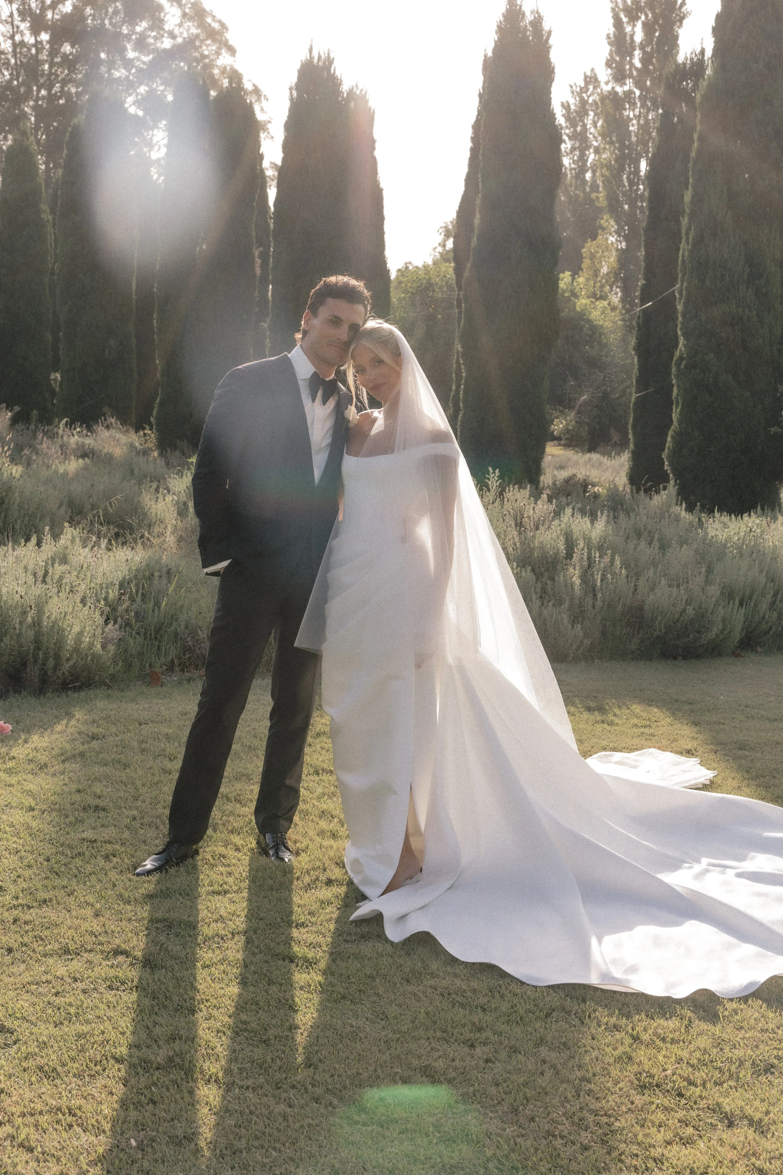 A newlywed couple standing outdoors on a grassy area with tall trees in the background, the groom wearing a black tuxedo and the bride in a white wedding dress with a long train and veil, sunlight streaming through the trees.