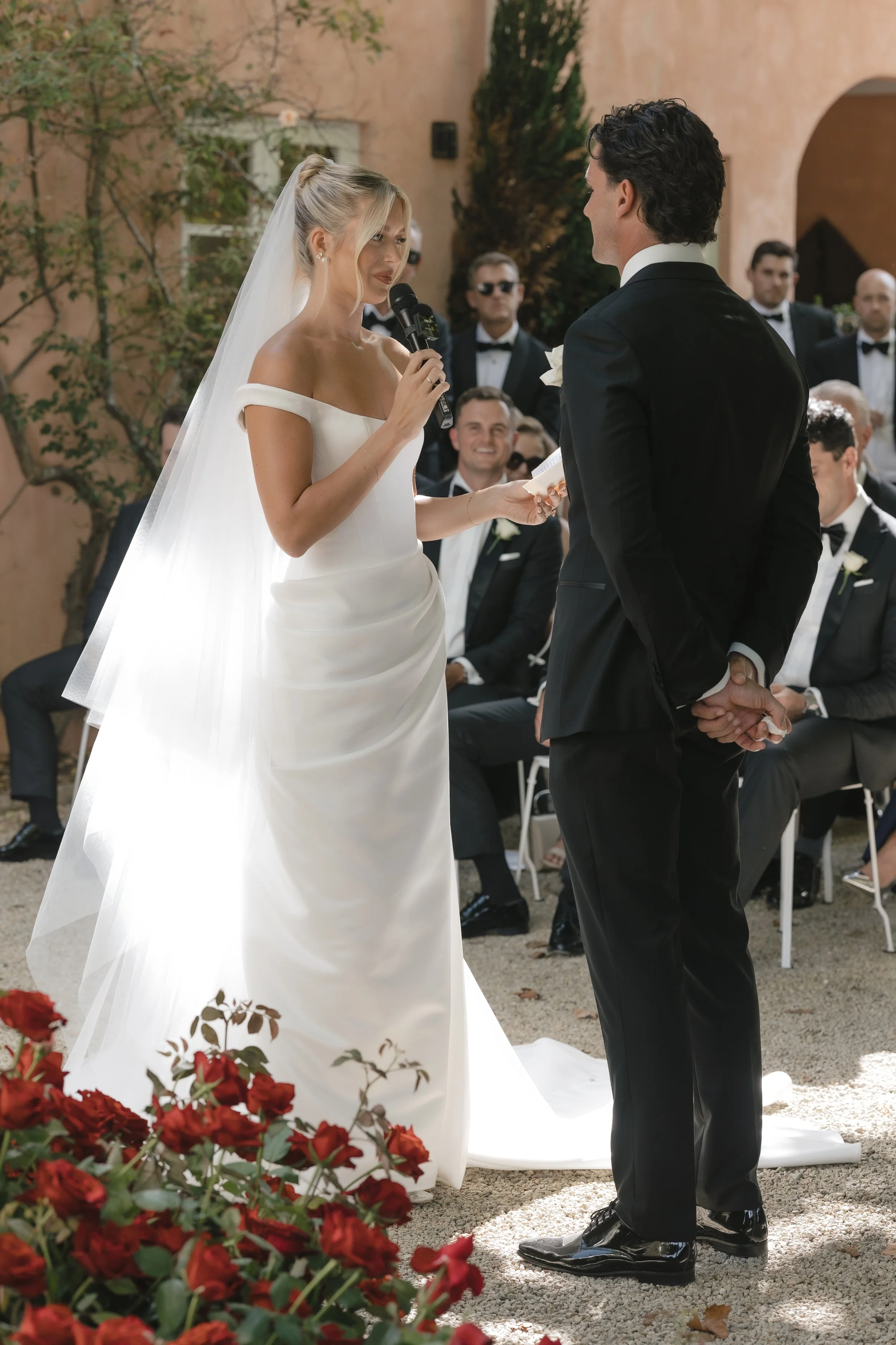 A bride and groom exchanging vows during their outdoor wedding ceremony, with guests seated in the background.