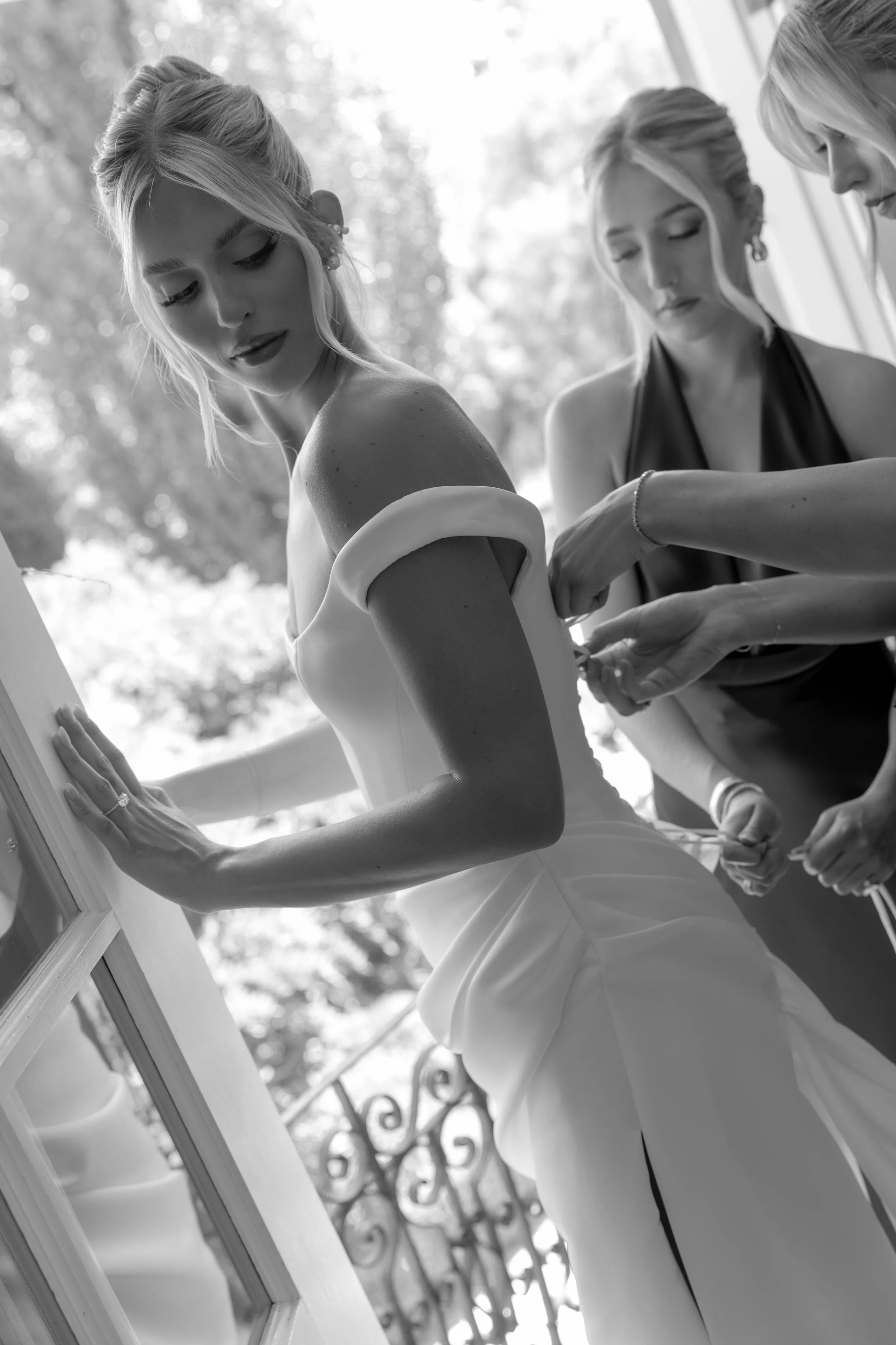 A bride in a wedding dress is being assisted with her dress by a woman as she gets ready for her wedding. The scene is outdoors, with trees and a decorative fence in the background.
