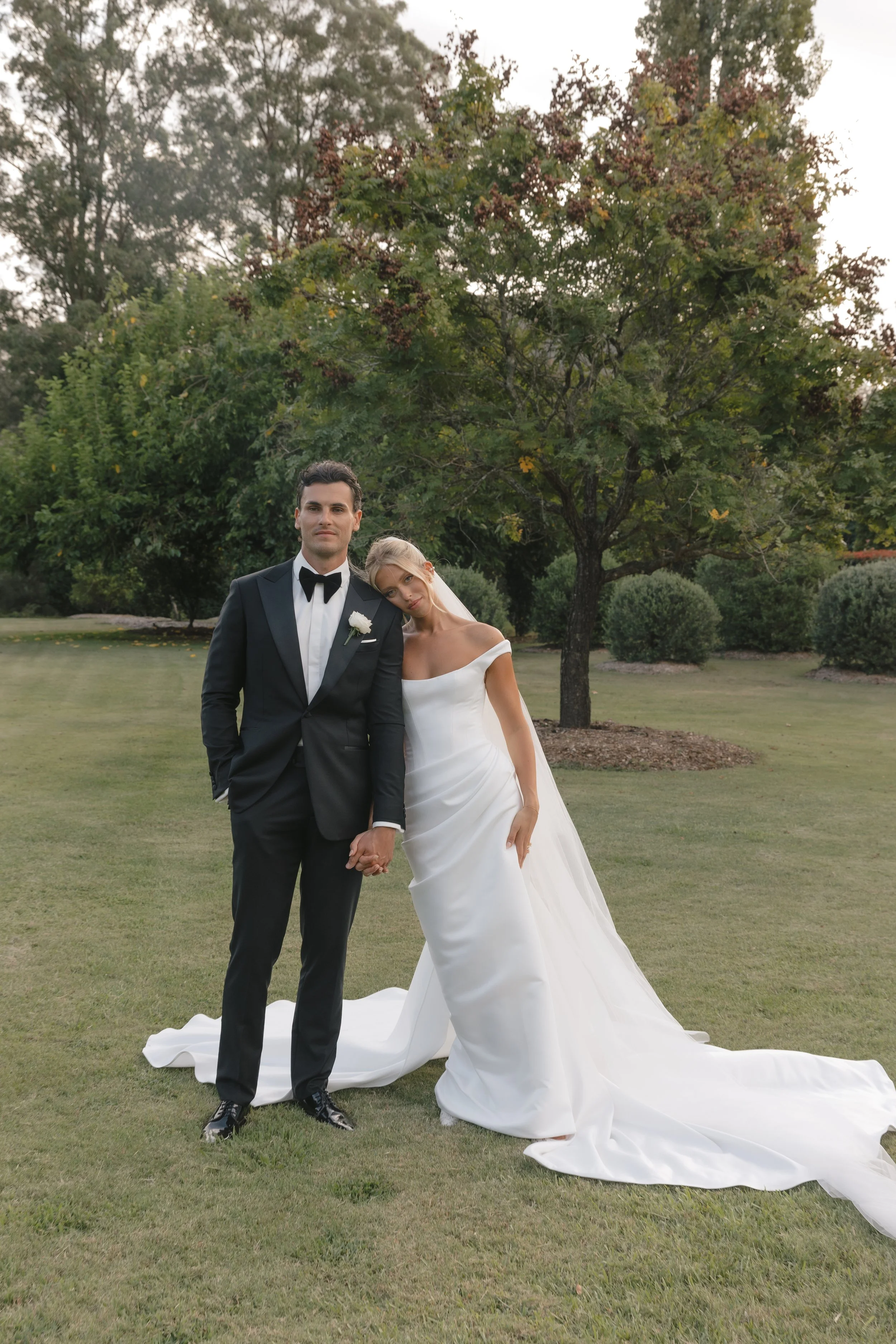 A bride and groom holding hands and posing for a photo outdoors. The groom is wearing a black tuxedo with a bow tie, and the bride is in a white wedding gown with off-the-shoulder sleeves. They are standing on a grassy area with trees in the background.