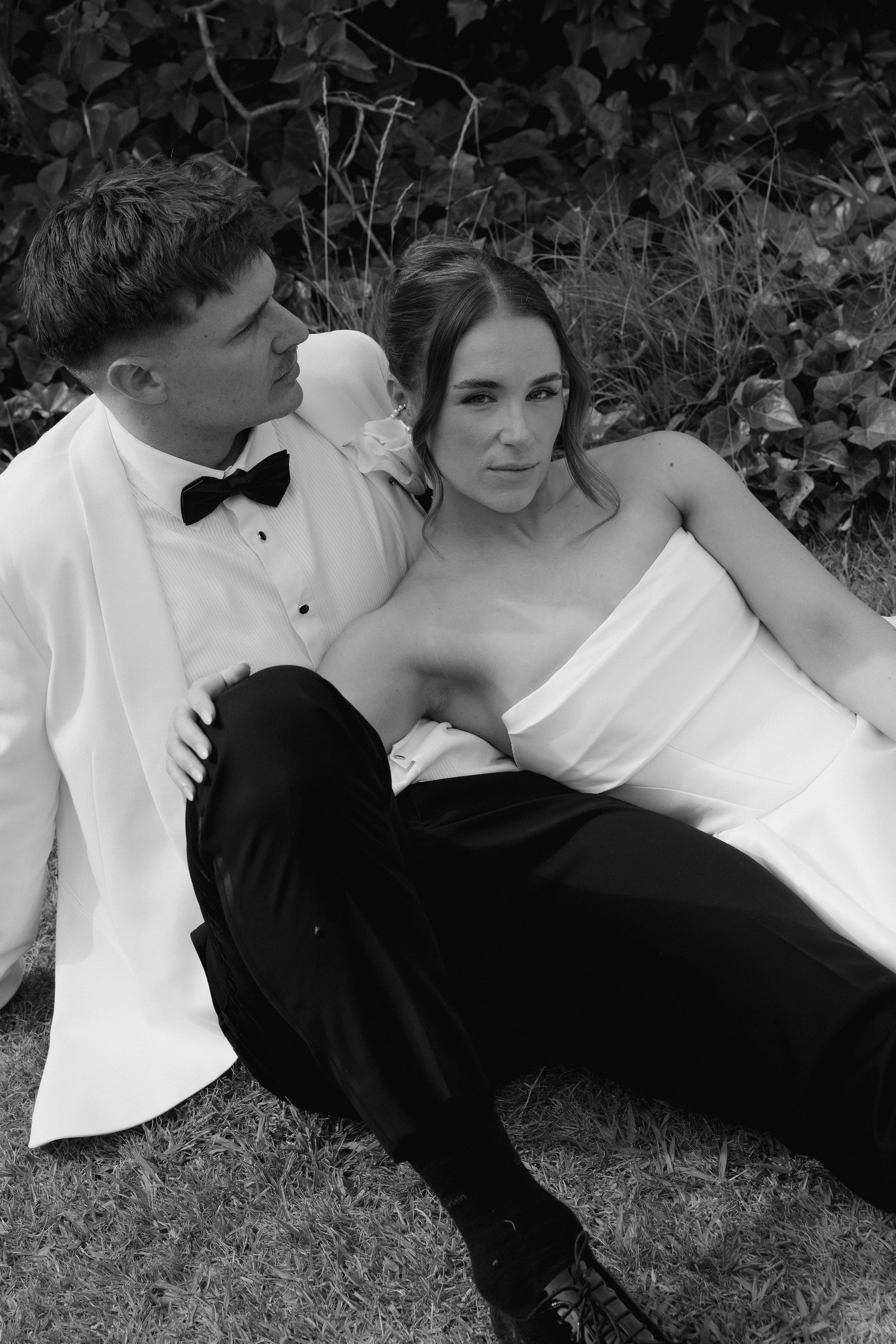 A black and white photo of a man in formal attire, including a tuxedo with a bow tie, and a woman in an elegant strapless dress, sitting on the grass with foliage in the background.