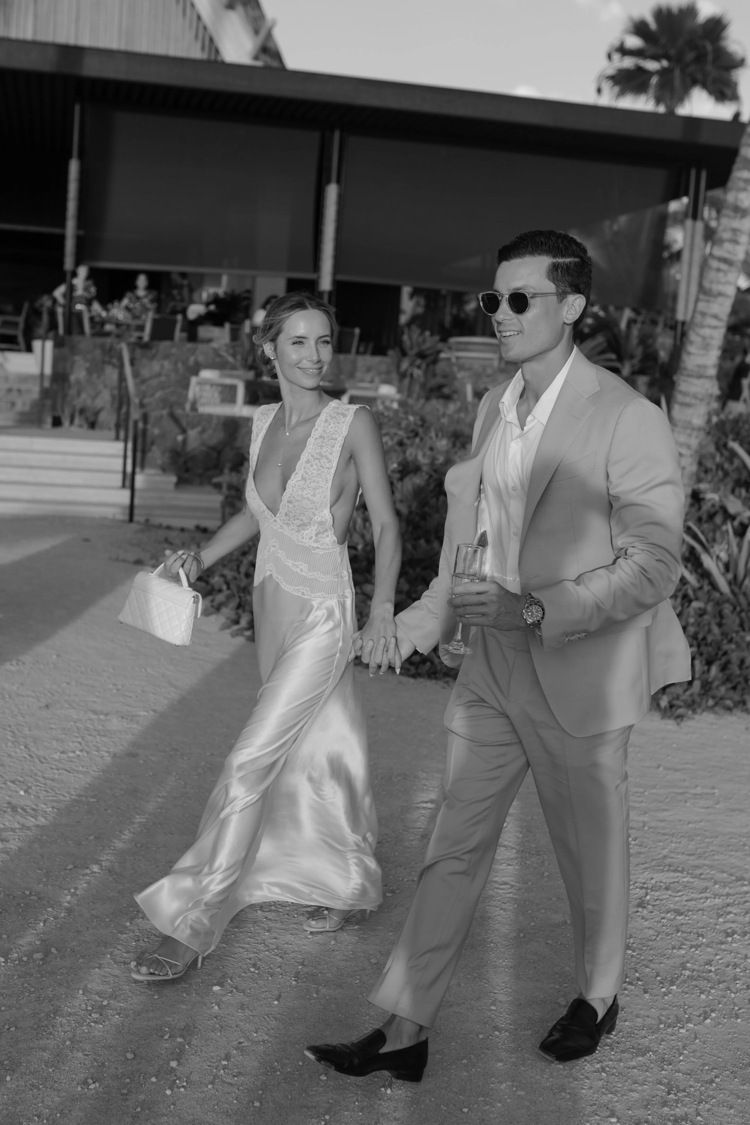 A woman and a man walking hand-in-hand outdoors, dressed in formal attire for a special occasion, with the woman holding a handbag and the man holding a glass, under a clear sky with palm trees and a modern building in the background.