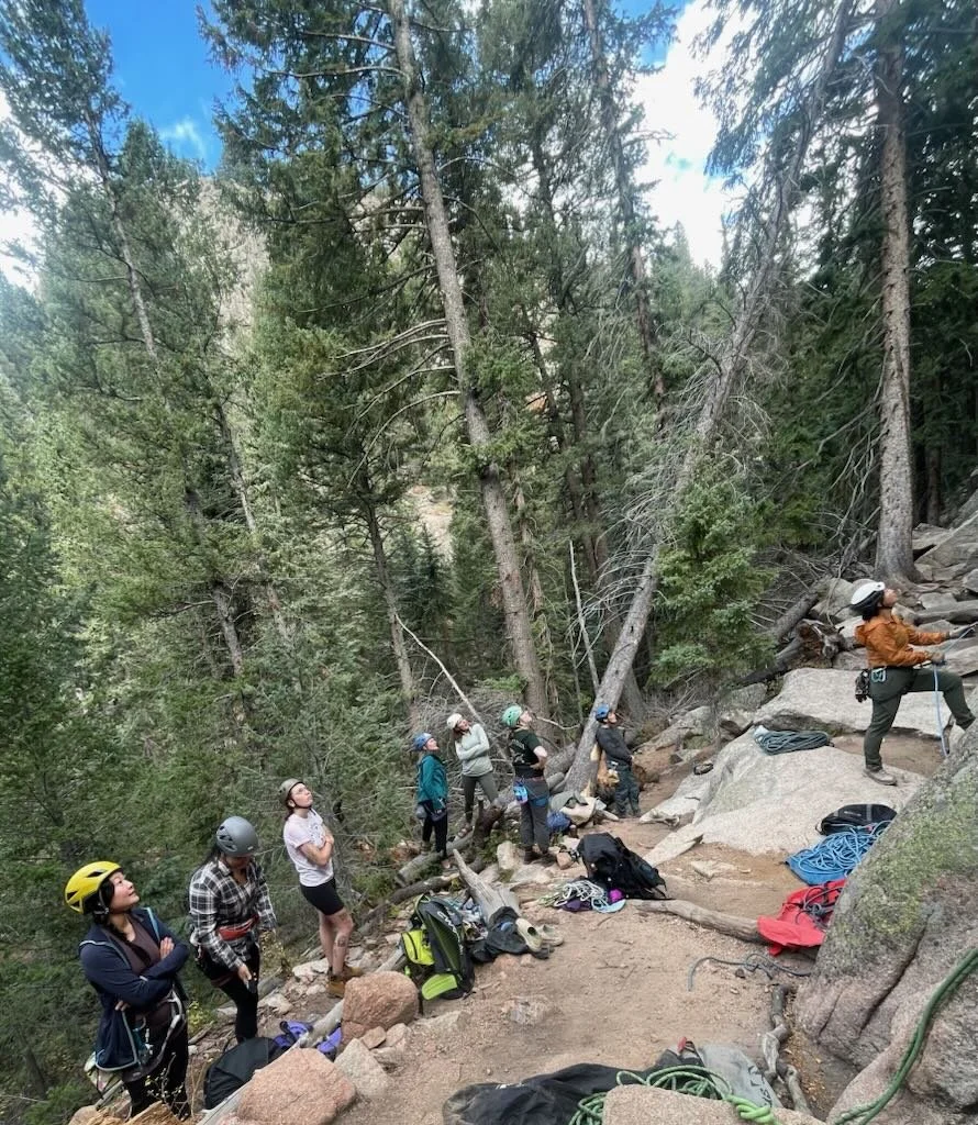 A group of people wearing helmets and outdoor gear standing on a rocky trail in a forest, preparing for rock climbing or rappelling.
