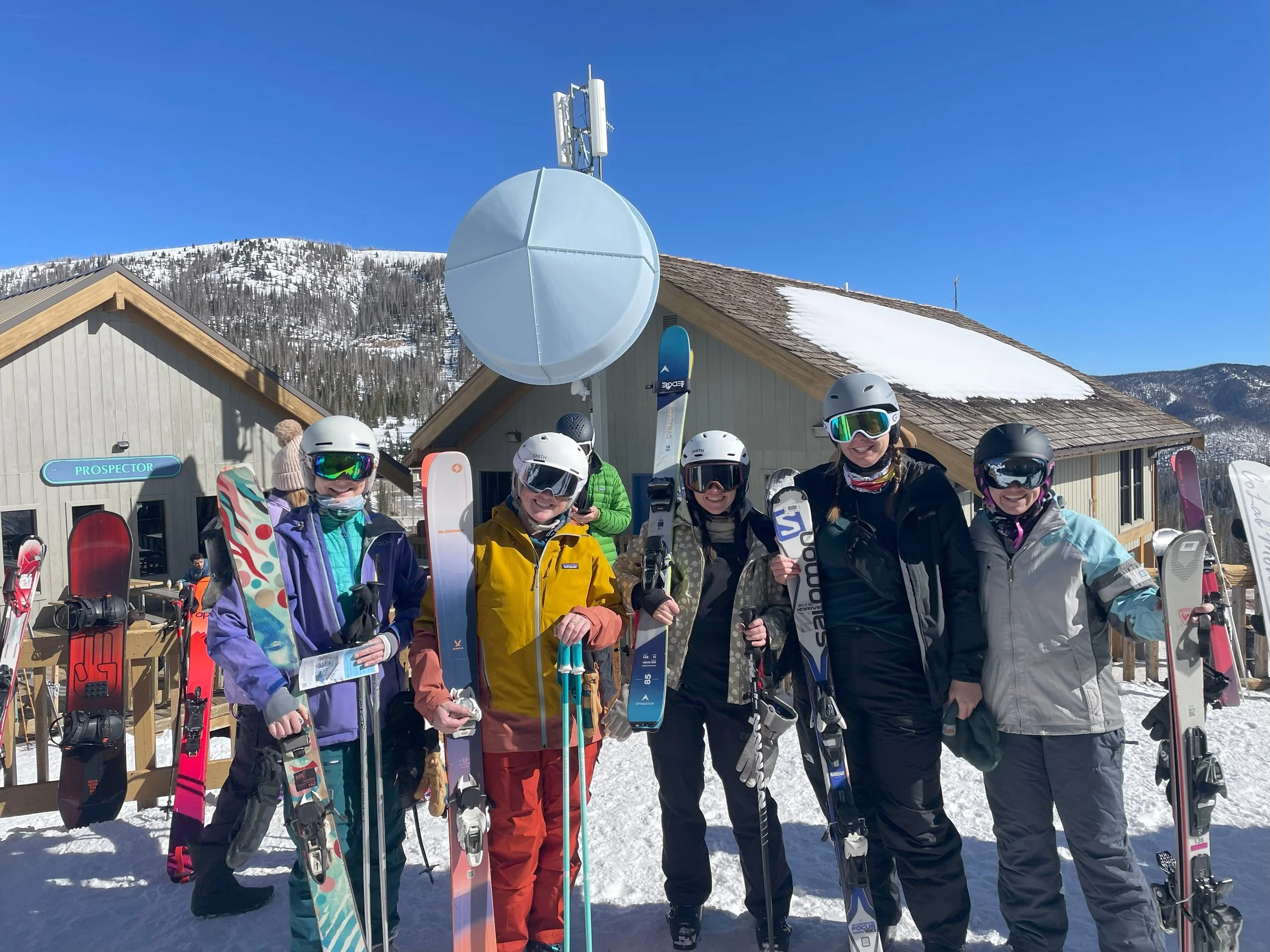 Group of six women dressed in winter ski gear, holding skis and standing in front of cabins at a snowy mountain resort with clear blue sky.