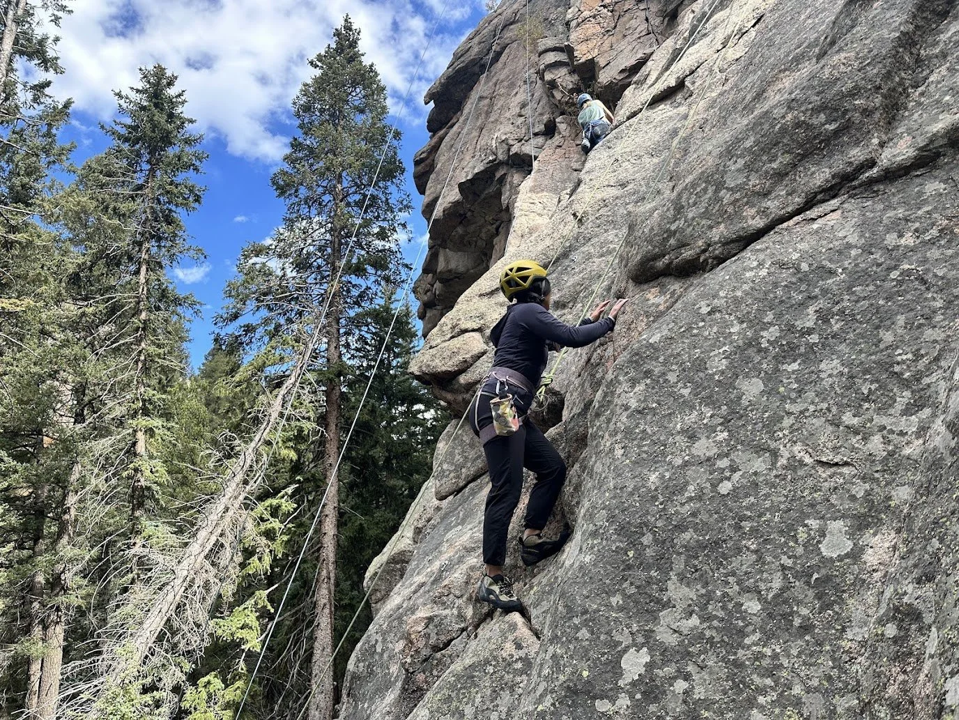A woman with a yellow helmet climbing a tall rock face using climbing gear, with a person above her also climbing, surrounded by trees under a partly cloudy sky.